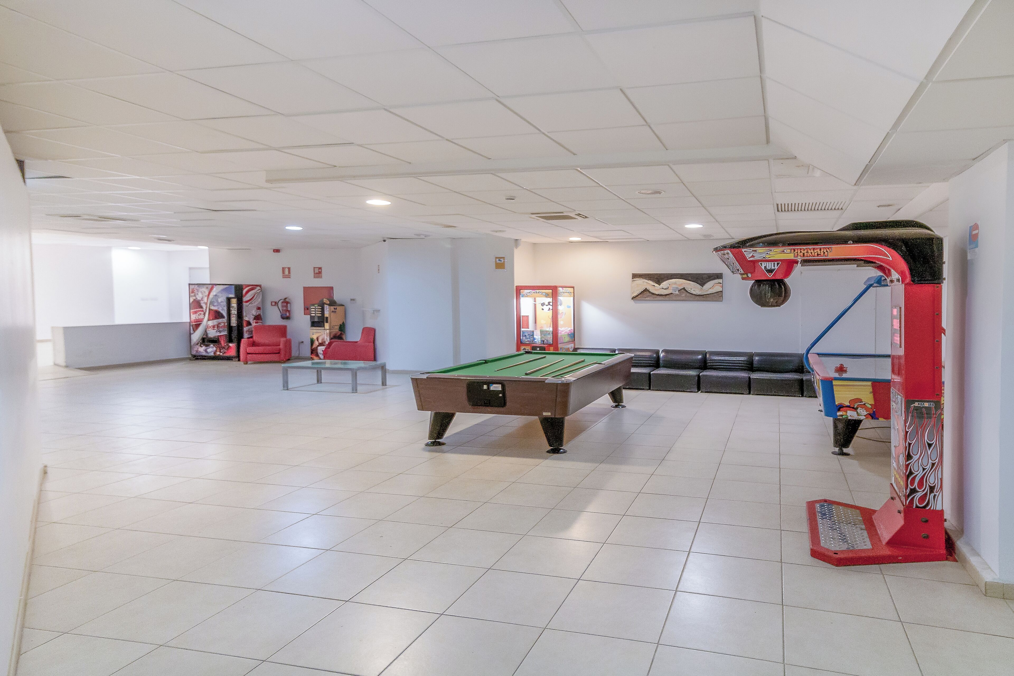 Een pooltafel in een ruime kamer met een plafondrooster, een rode en blauwe speelautomaat en een zithoek met rode stoelen.