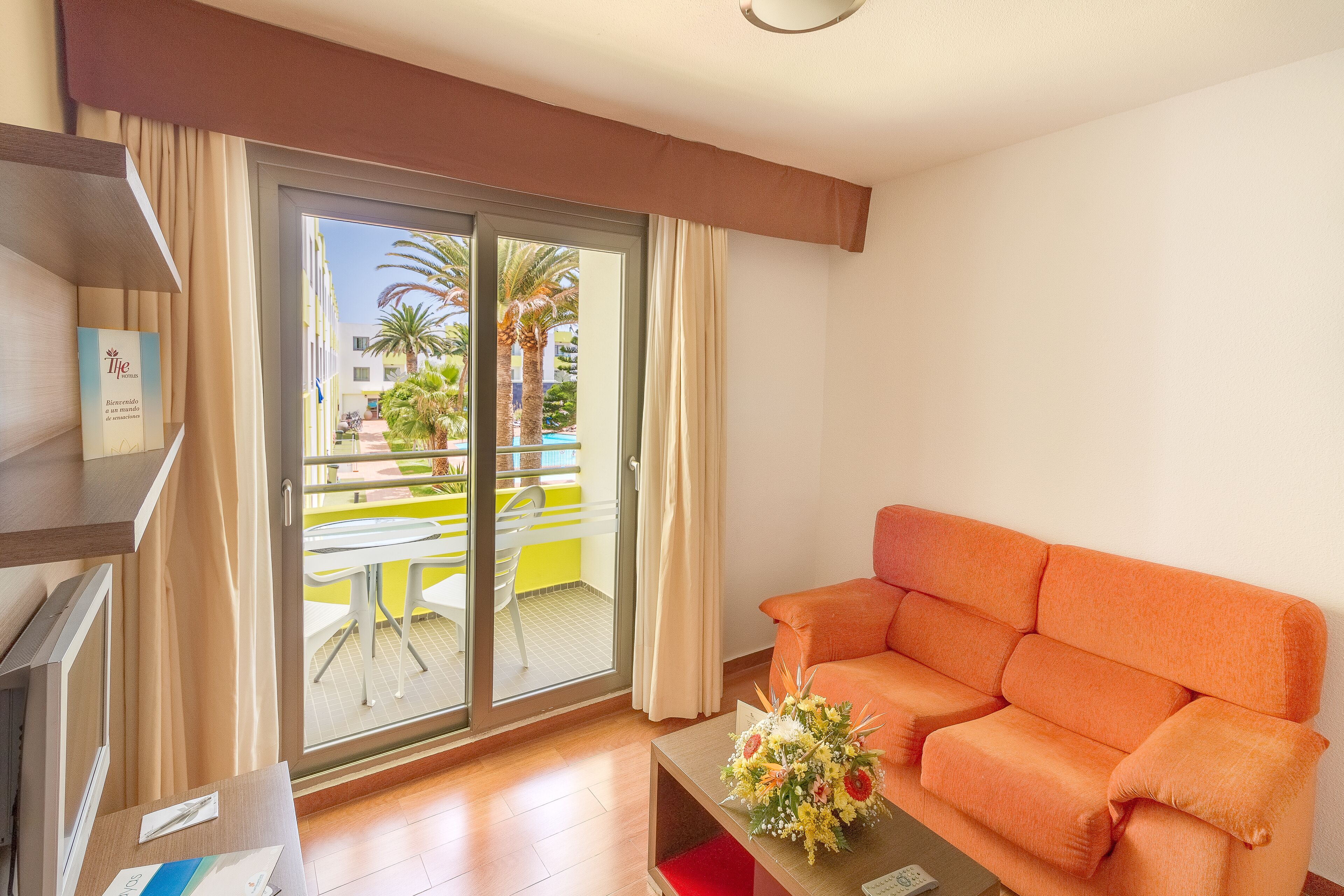 A hotel room with a sliding glass door to a balcony, a sofa, a small table with a flower arrangement, and a TV on a stand.