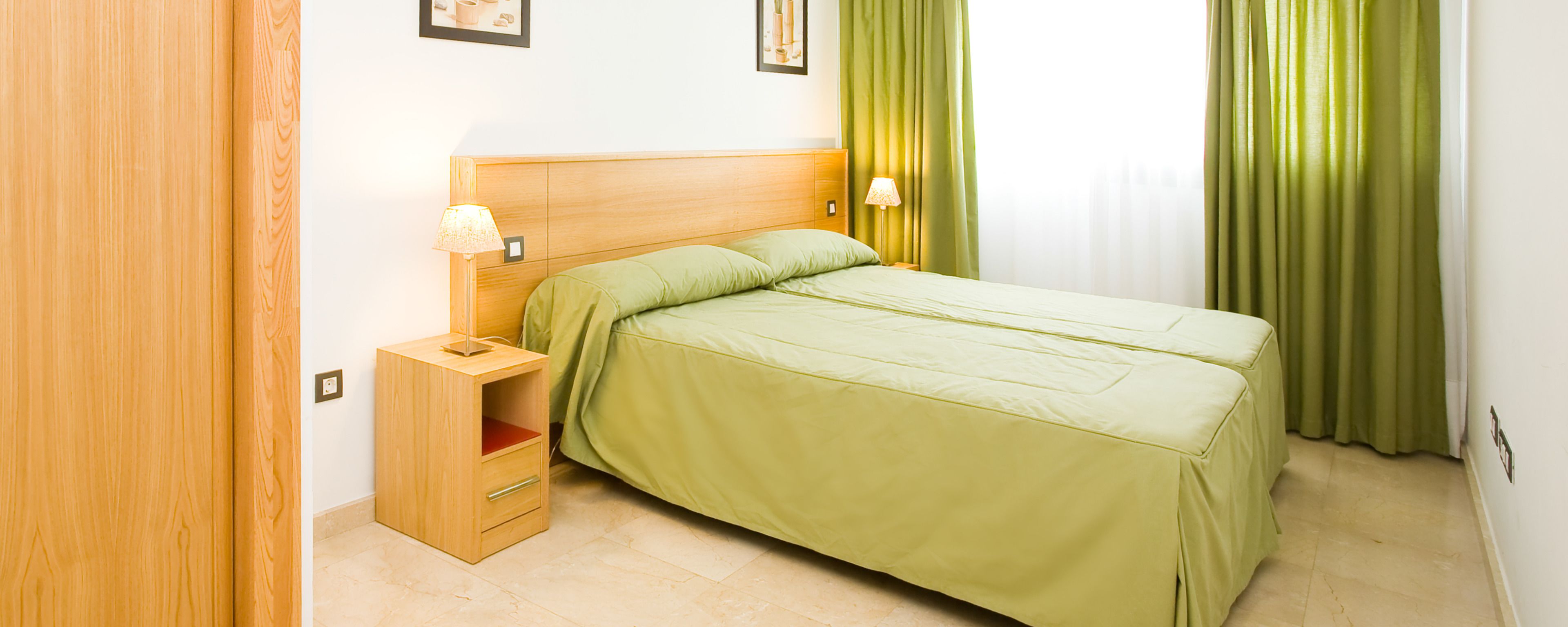 Ein ordentlich eingerichtetes Schlafzimmer mit einem Holzkopfteil, einem Einzelbett mit grüner Bettwäsche, einem Nachttisch mit Lampe und einem grünen Vorhang.