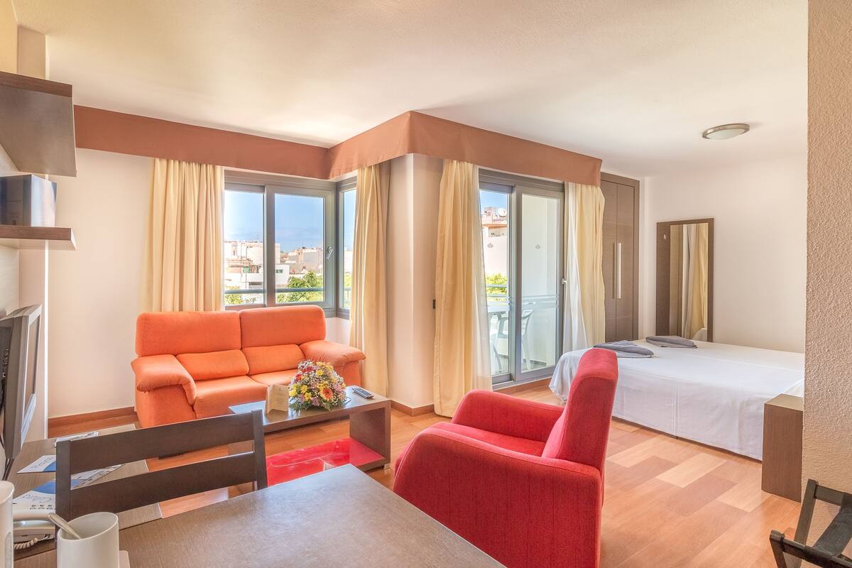 Une chambre d’hôtel moderne équipée d’un canapé, d’un fauteuil, d’un lit, d’une télévision et d’un balcon avec des rideaux.