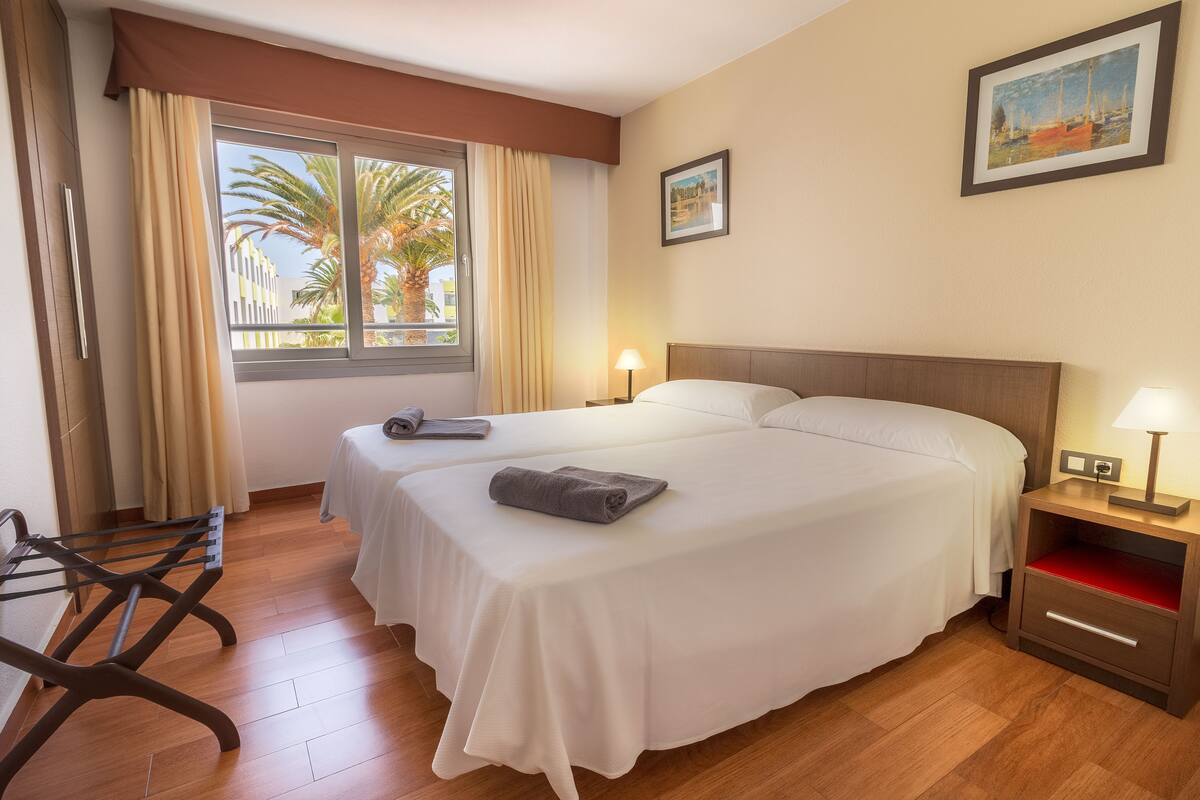 Une chambre d’hôtel avec deux lits, un plancher en bois, une fenêtre avec des rideaux, une table de chevet avec une lampe et un tableau accroché au mur.