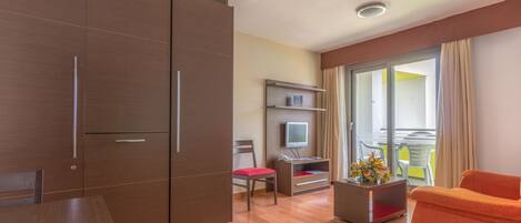 A modern hotel room with a wooden wardrobe, a dining table, a sofa, a TV, and a balcony with a view.