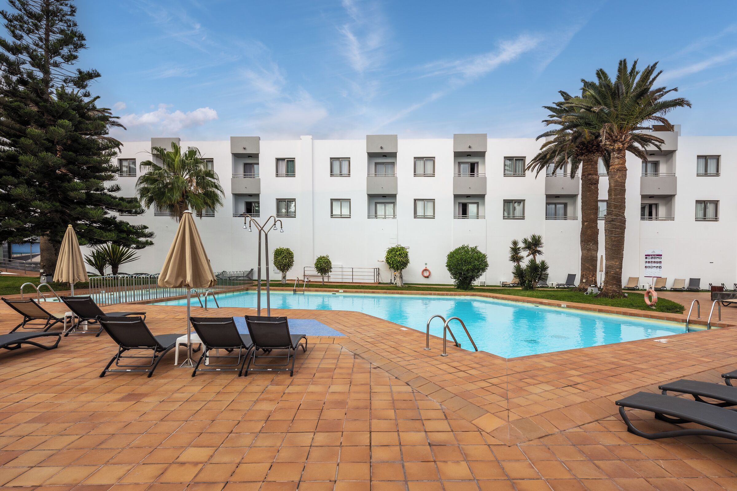 A swimming pool area with lounge chairs and umbrellas, surrounded by a building with multiple balconies and palm trees.