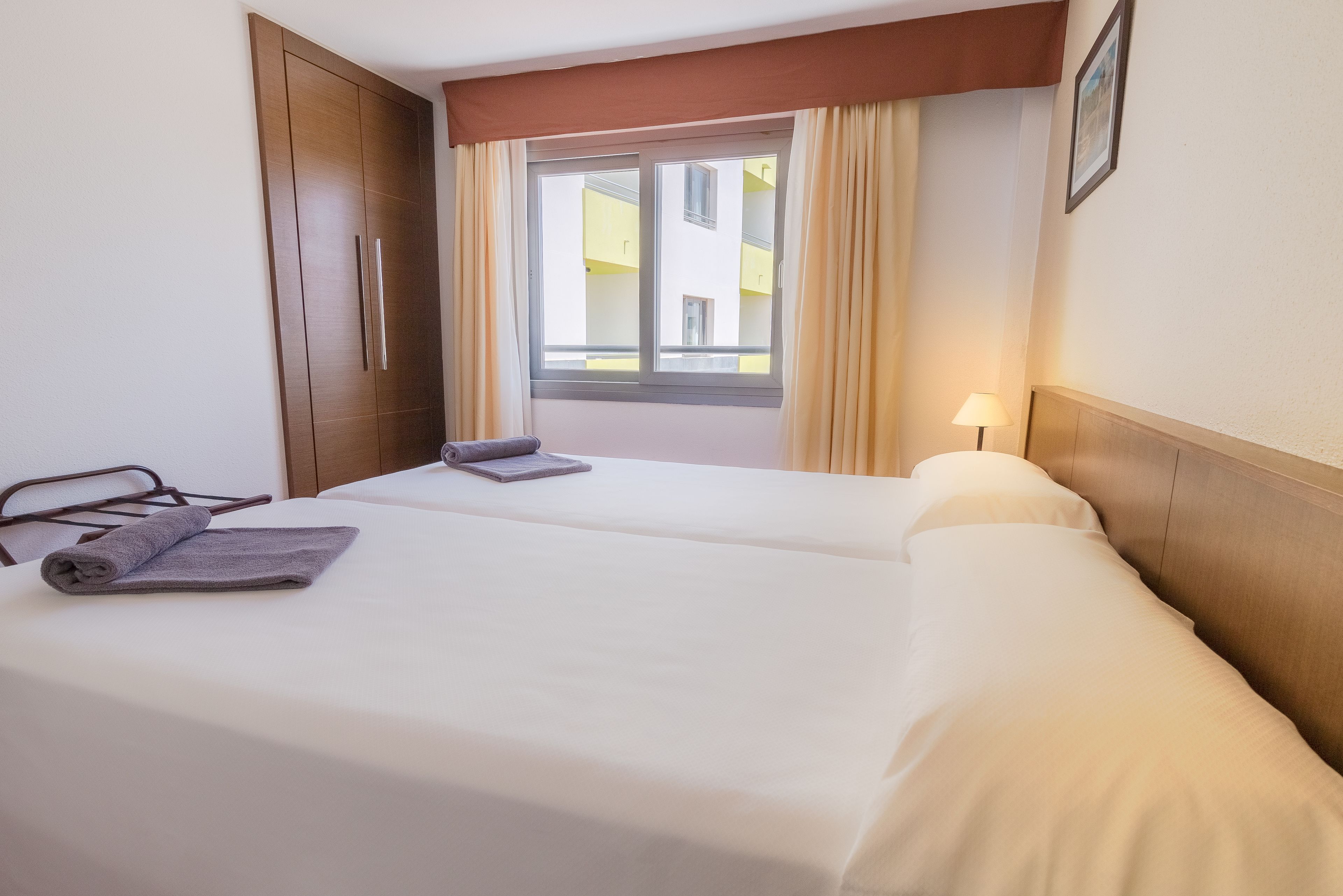 A hotel room with two beds, a window with curtains, a lamp, and a wooden wardrobe.