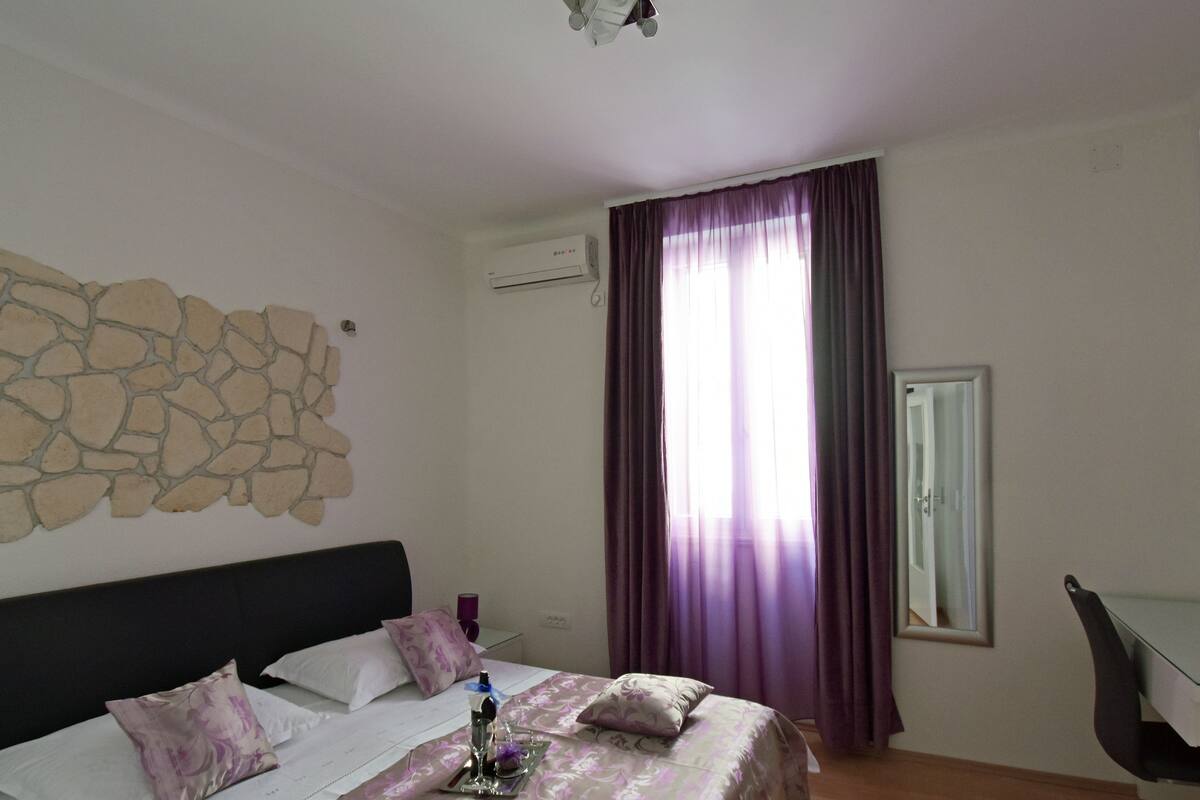 Ein Hotelzimmer mit Steinwand, einem Bett mit floralem Überwurf, einem Schreibtisch mit Stuhl und einem Fenster mit violetten Vorhängen.