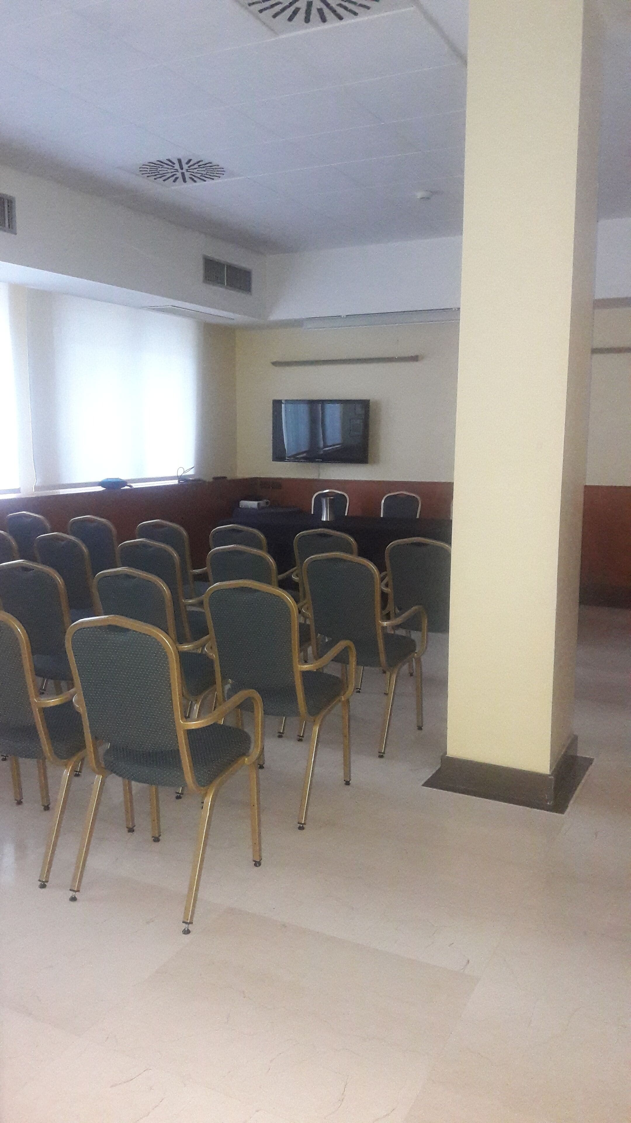 Un salón de conferencias con filas de sillas, un televisor en la pared y un pilar.