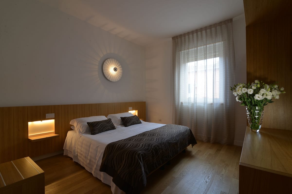 Une chambre à coucher comprenant un lit, des tables de chevet, une lampe fixée au mur, une fenêtre avec des rideaux et un vase avec des fleurs.