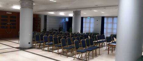 A conference room with rows of blue chairs, a large table, and a high ceiling with recessed lighting.