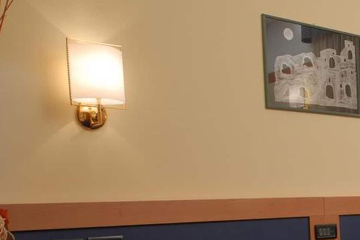 Une chambre d’hôtel avec un lit, une table de chevet, une lampe, un tableau encadré au mur et une petite étagère sur laquelle est posé un vase de fleurs.