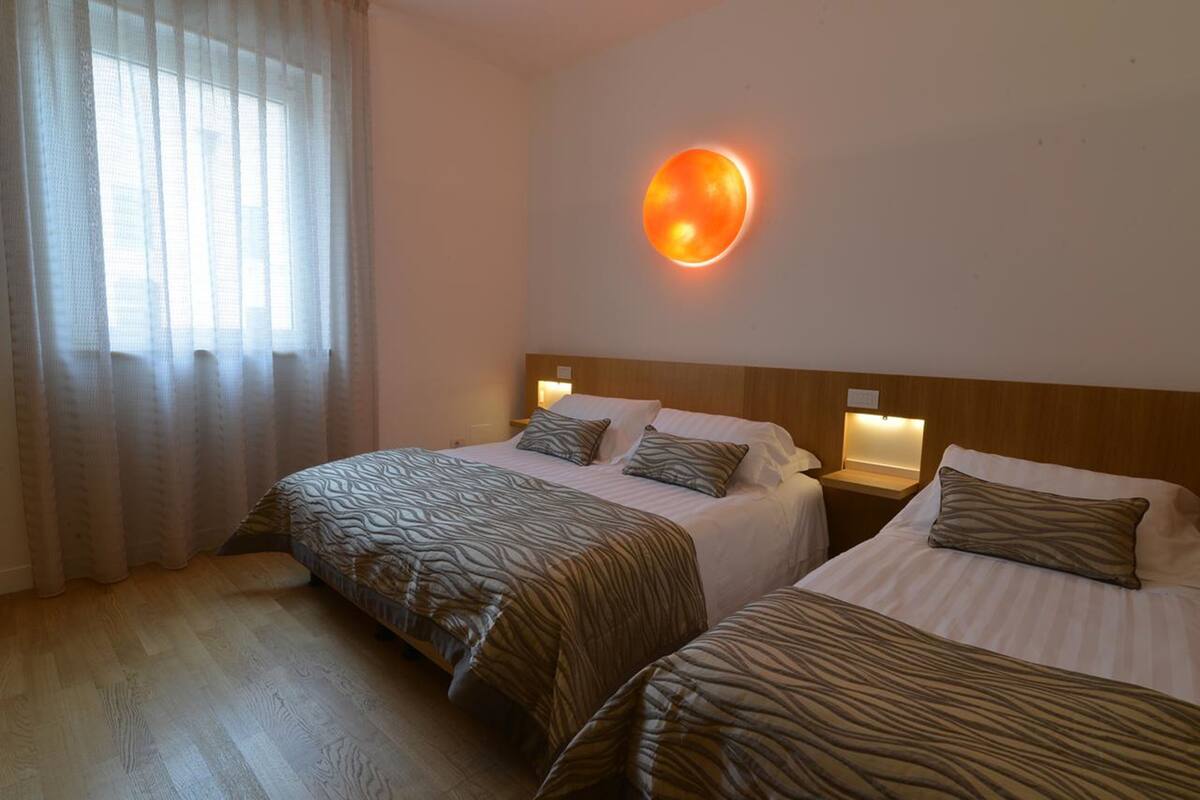 Une chambre d’hôtel avec deux lits, une grande fenêtre avec des rideaux légers et un luminaire fixé au mur.