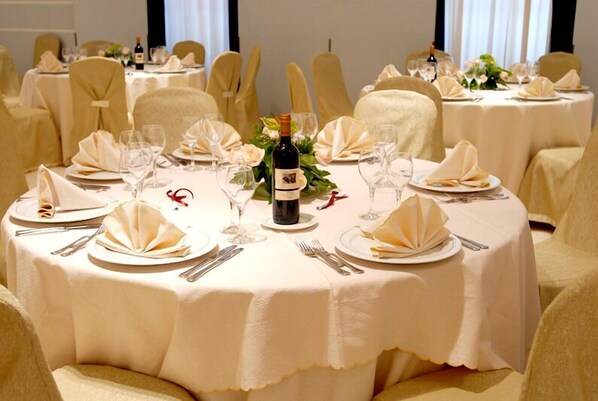 Un salón de banquetes con mesas redondas dispuestas para un evento formal, cada una con manteles blancos, servilletas dobladas y botellas de vino.