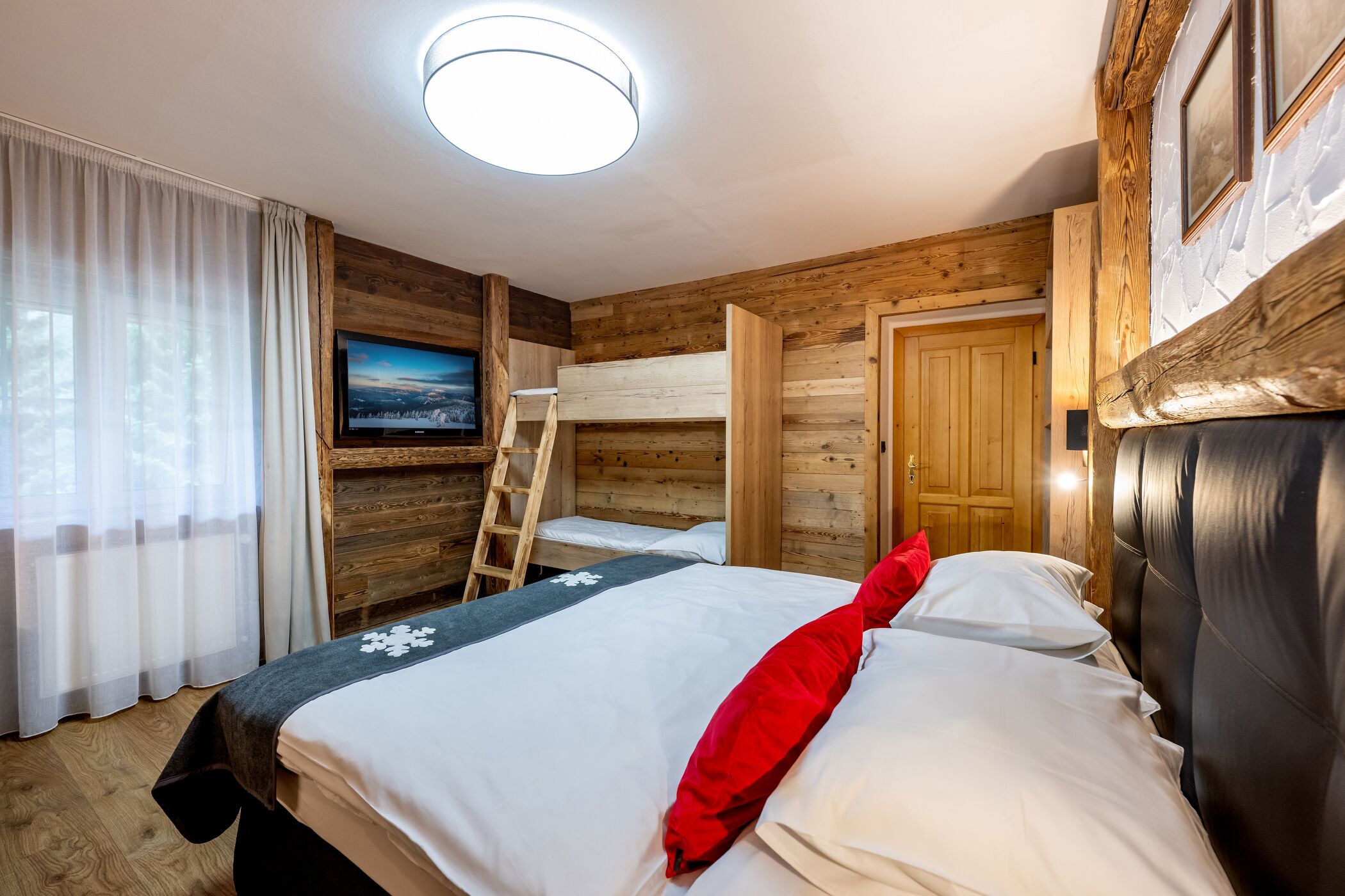 A cozy room with a bunk bed, a wooden ladder, a flat-screen TV, and a large window with curtains.