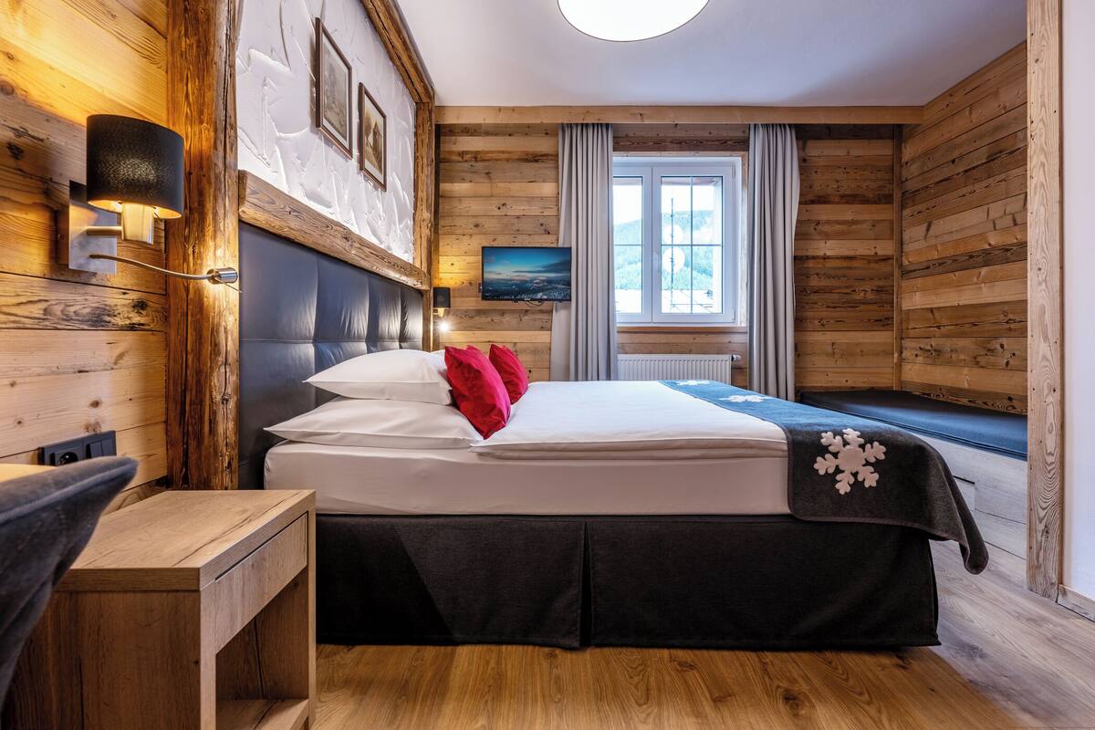 Una habitación con paneles de madera, una cama, una mesita de noche, una lámpara de pared, una ventana con cortinas y un cuadro en la pared.