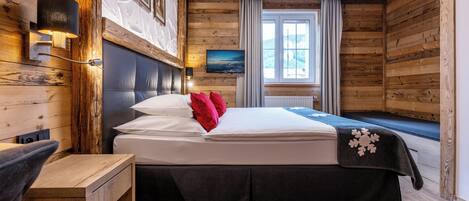 A wooden-paneled room with a bed, a nightstand, a wall-mounted lamp, a window with curtains, and a painting on the wall.