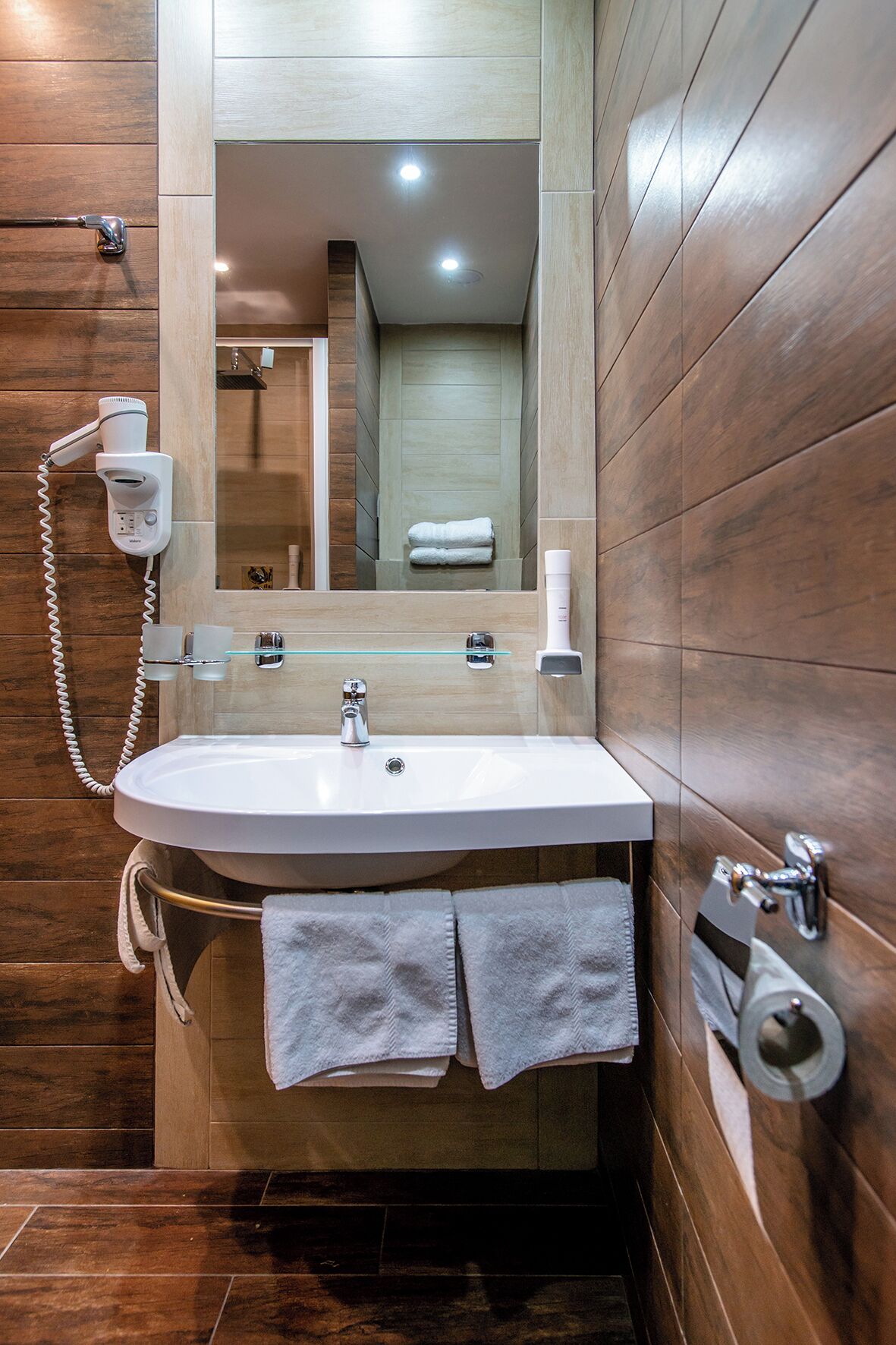 A modern bathroom with a wooden floor, a white sink, a glass shelf, a towel rack, and a hairdryer.