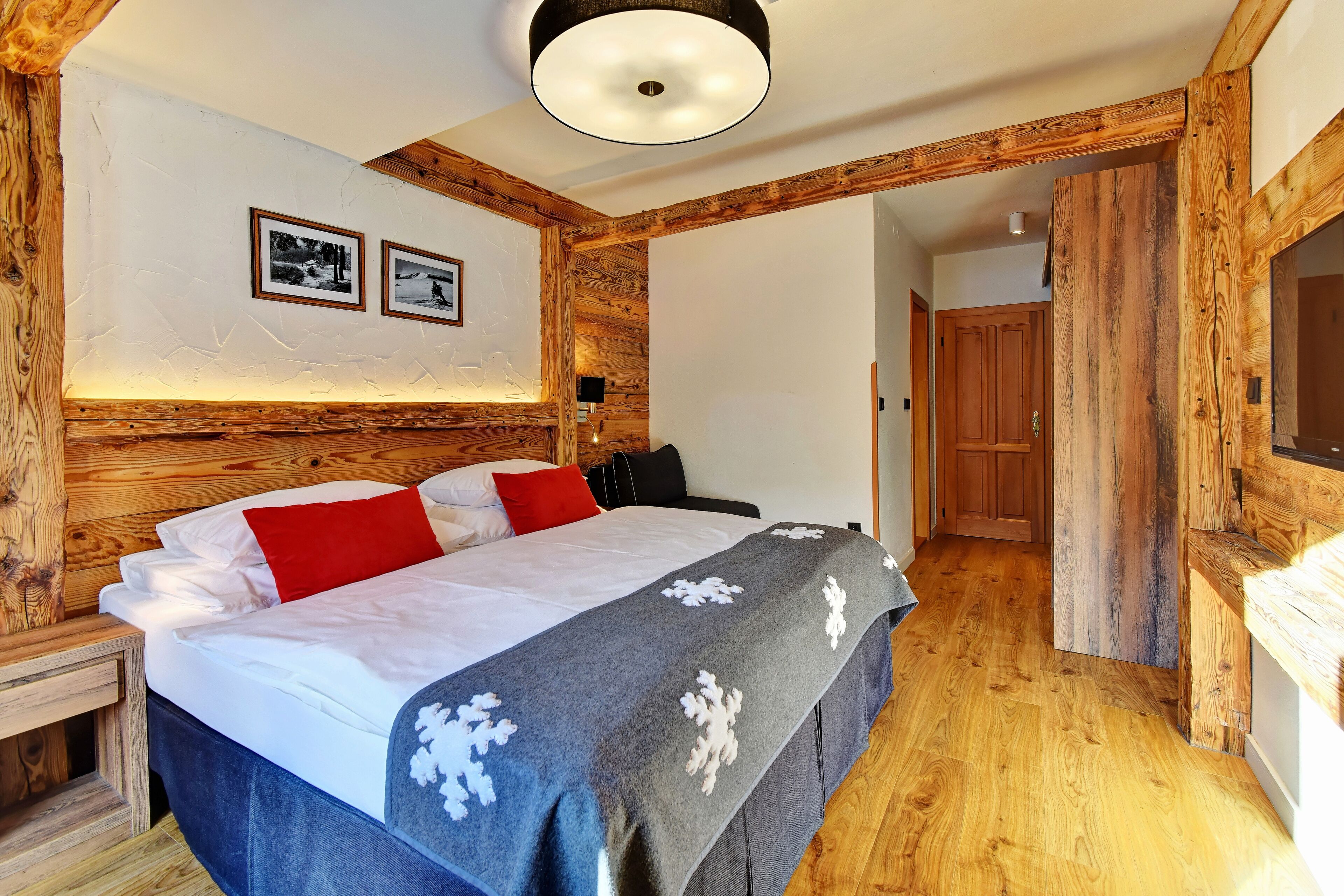 Un dormitorio con cabecera de madera, cama con ropa blanca y almohadas rojas, manta gris con motivos de copos de nieve blancos, y dos cuadros en la pared.