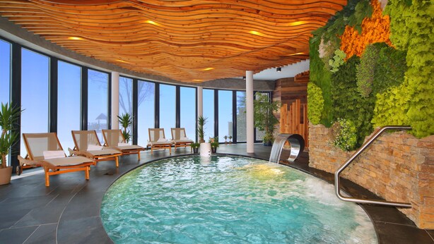 Una piscina interior moderna con sillones de descanso, techo de madera y pared verde con plantas.