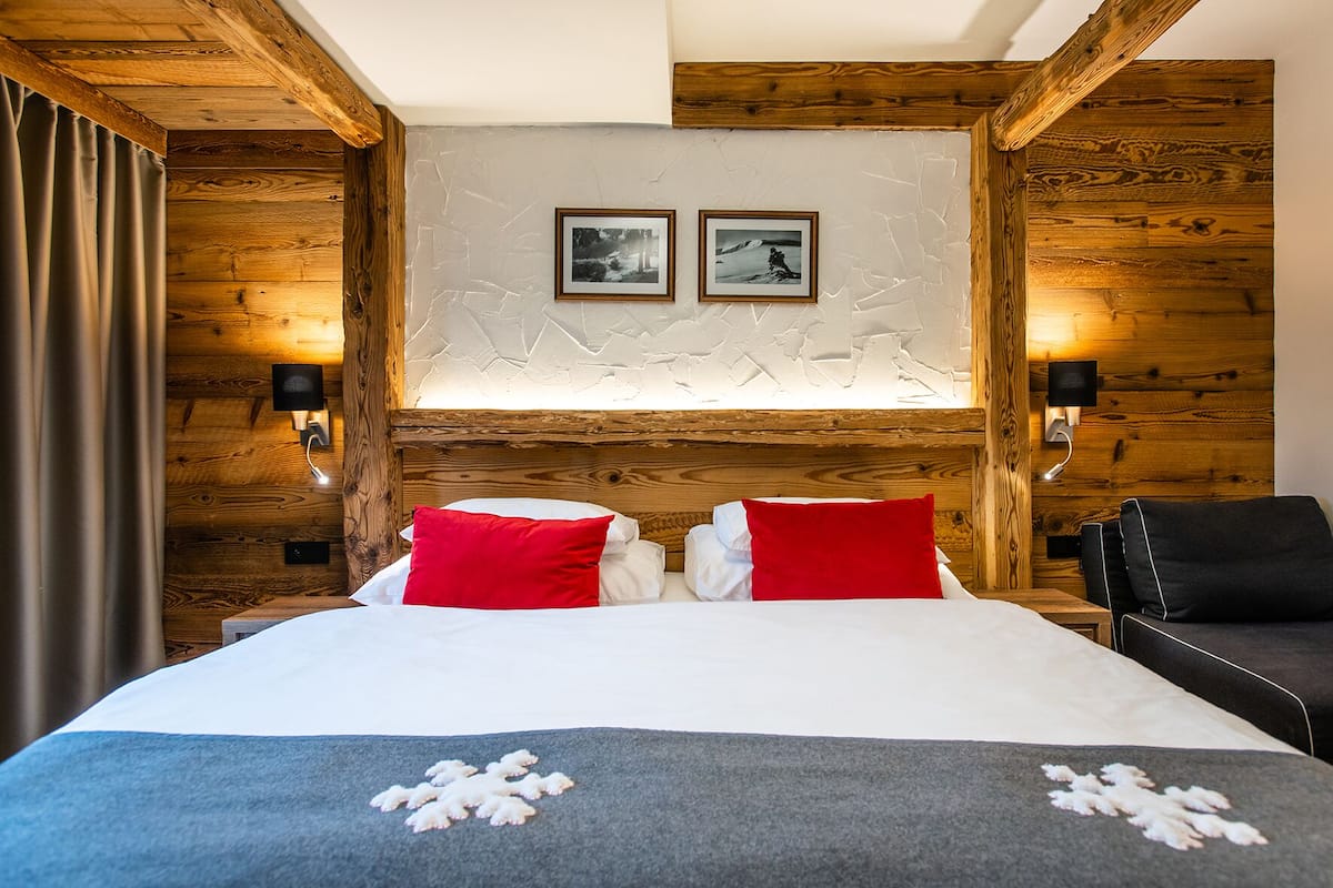 Habitación de hotel con cabecera de madera, dos cuadros enmarcados en la pared, cama con almohadas rojas y blancas, y manta gris con diseño de copo de nieve.