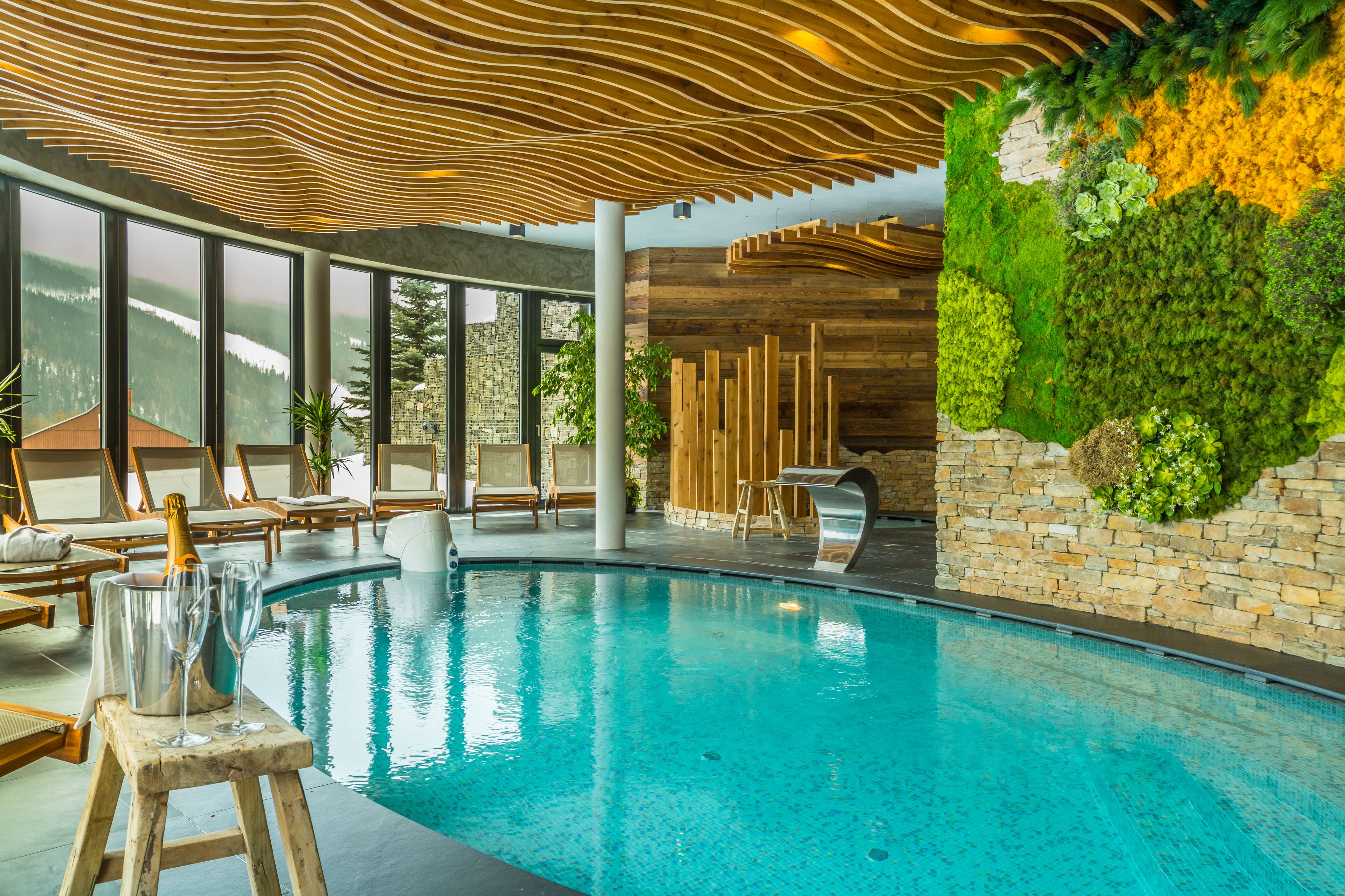 A modern indoor pool area with lounge chairs, a wooden ceiling, and a green wall with plants.