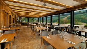 Un restaurante con vigas de madera, suelo de piedra y amplios ventanales que dan a una ladera boscosa.