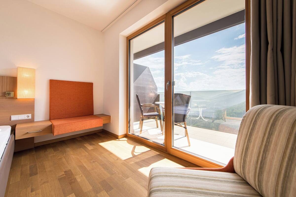 Ein modernes Hotelzimmer mit einer großen Schiebeglasdoor zum Balkon, einem Bett, einem Nachttisch und einem Sessel.