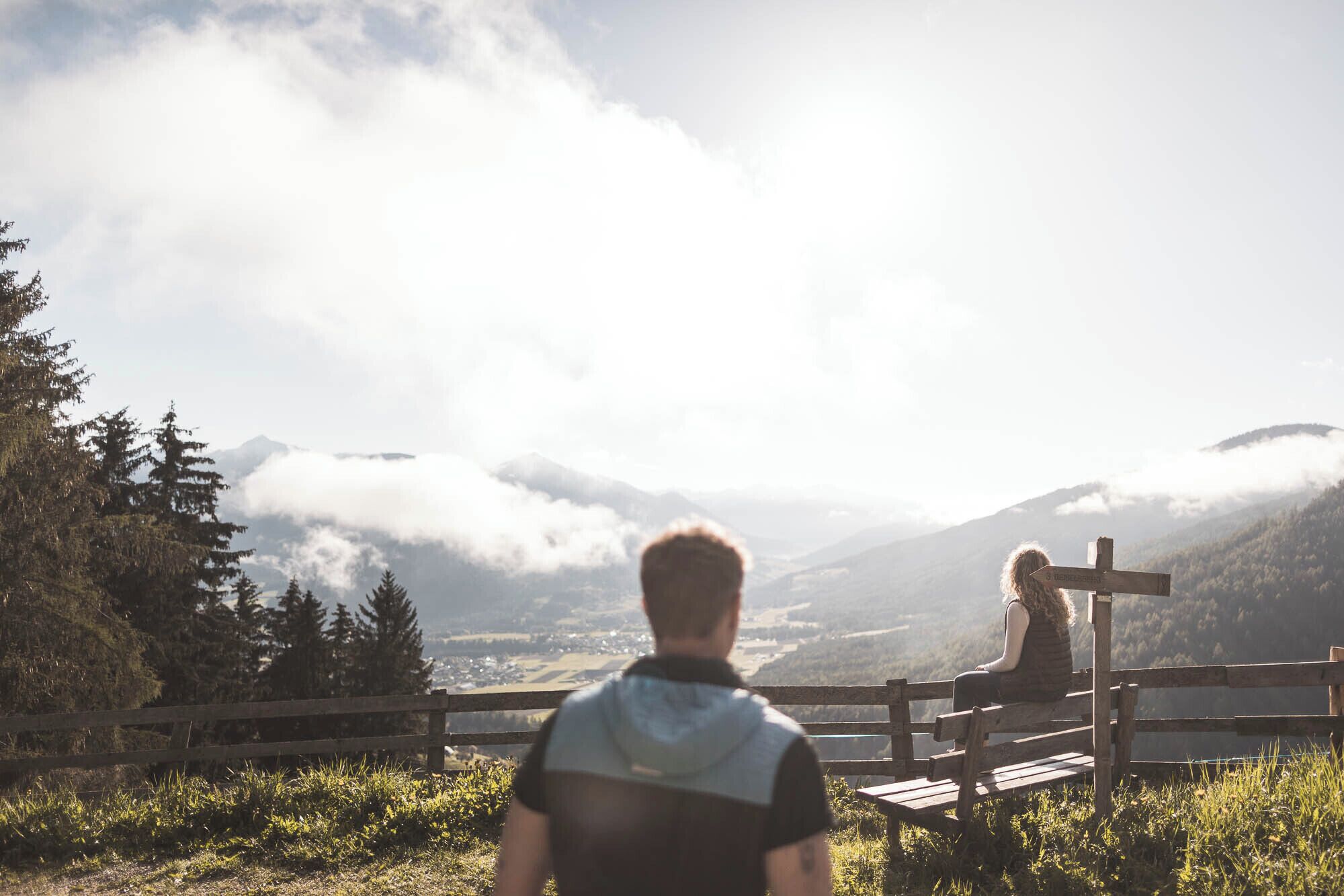 Eine Person sitzt auf einer Bank und blickt auf eine gebirgige Landschaft. Im Vordergrund steht ein hölzernes Wegweiser.