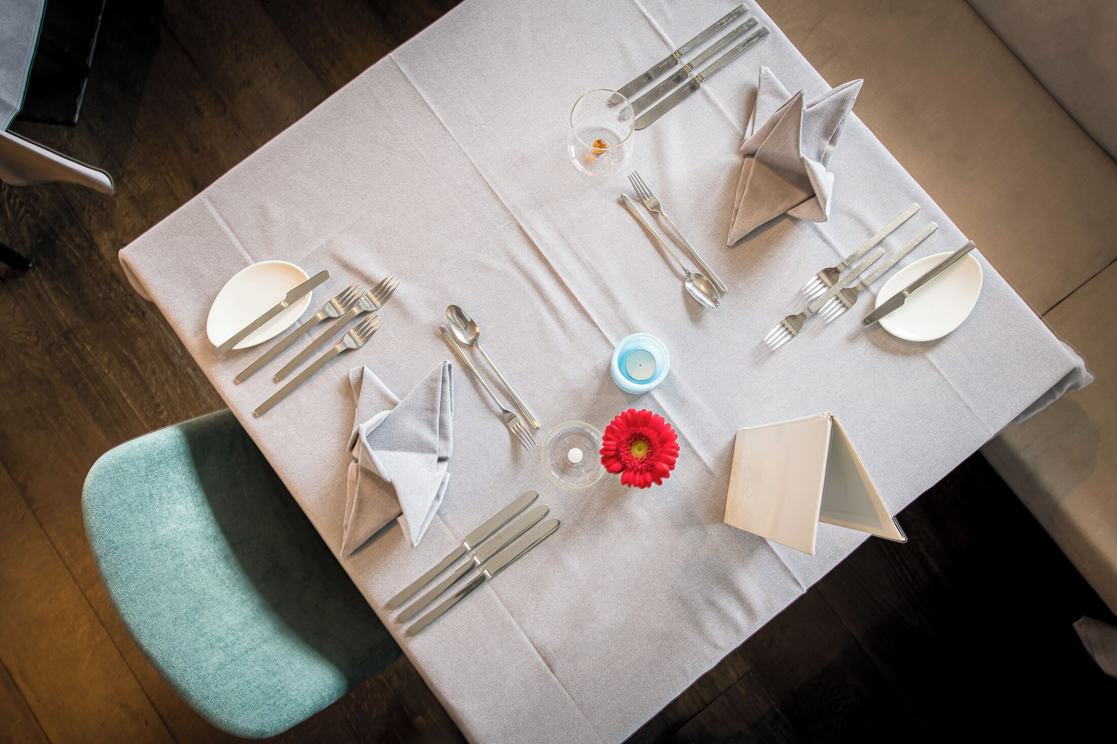 Una mesa de comedor elegantemente dispuesta, con mantel blanco, cubiertos, platos y un centro de mesa con una flor roja.