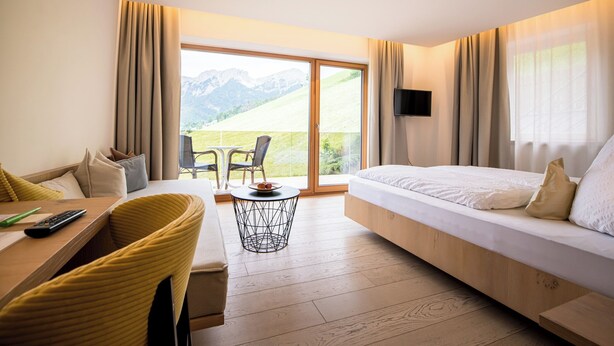 A modern hotel room with a bed, a dining area, and a balcony with a mountain view. - Kronplatz Resort Hotel Kristall (Valdaora)