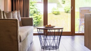 A living room with a sofa, a round table with a wireframe base, and a view of a garden through a sliding glass door. - Kronplatz Resort Hotel Kristall (Valdaora)