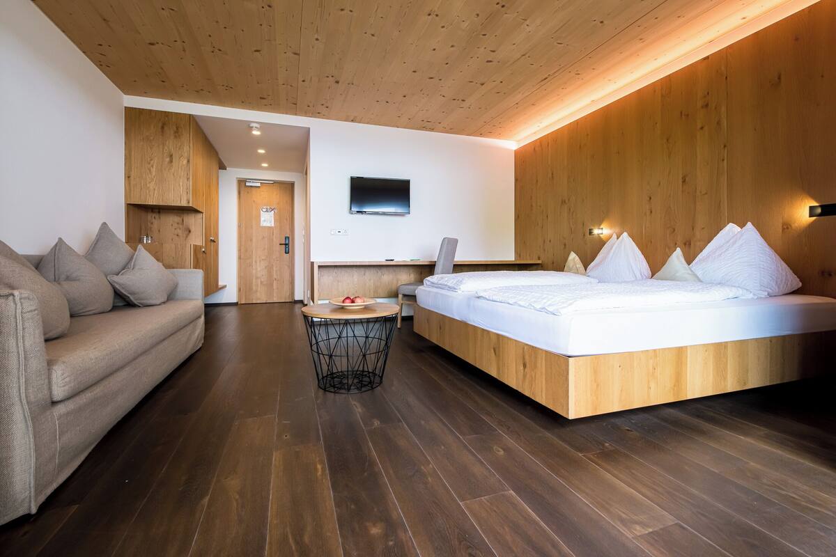 Ein modernes Schlafzimmer mit Holzboden, einem Bett mit weißer Bettwäsche, einer Couch mit Kissen und einem kleinen runden Tisch mit einer Obstschale.