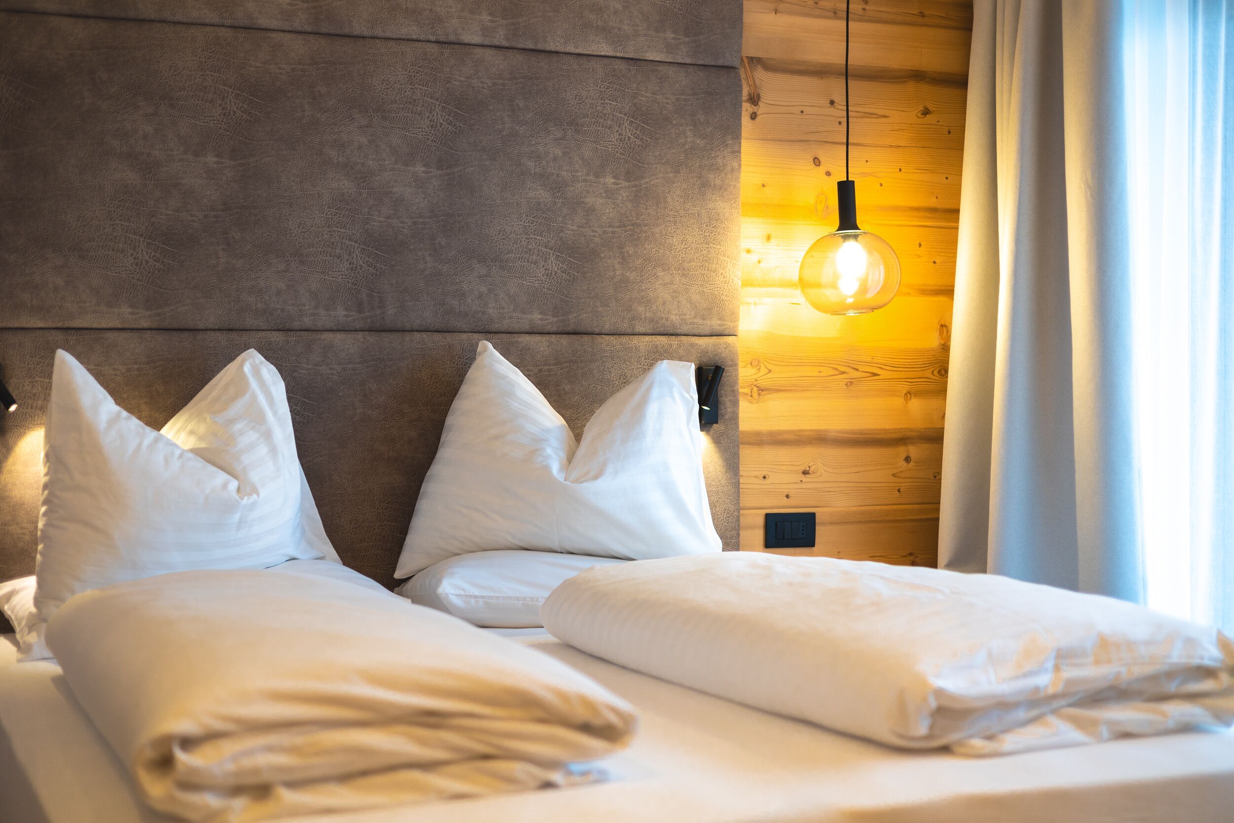 Una cama bien hecha con sábanas y almohadas blancas, un cabecero de madera y una lámpara fijada a la pared.