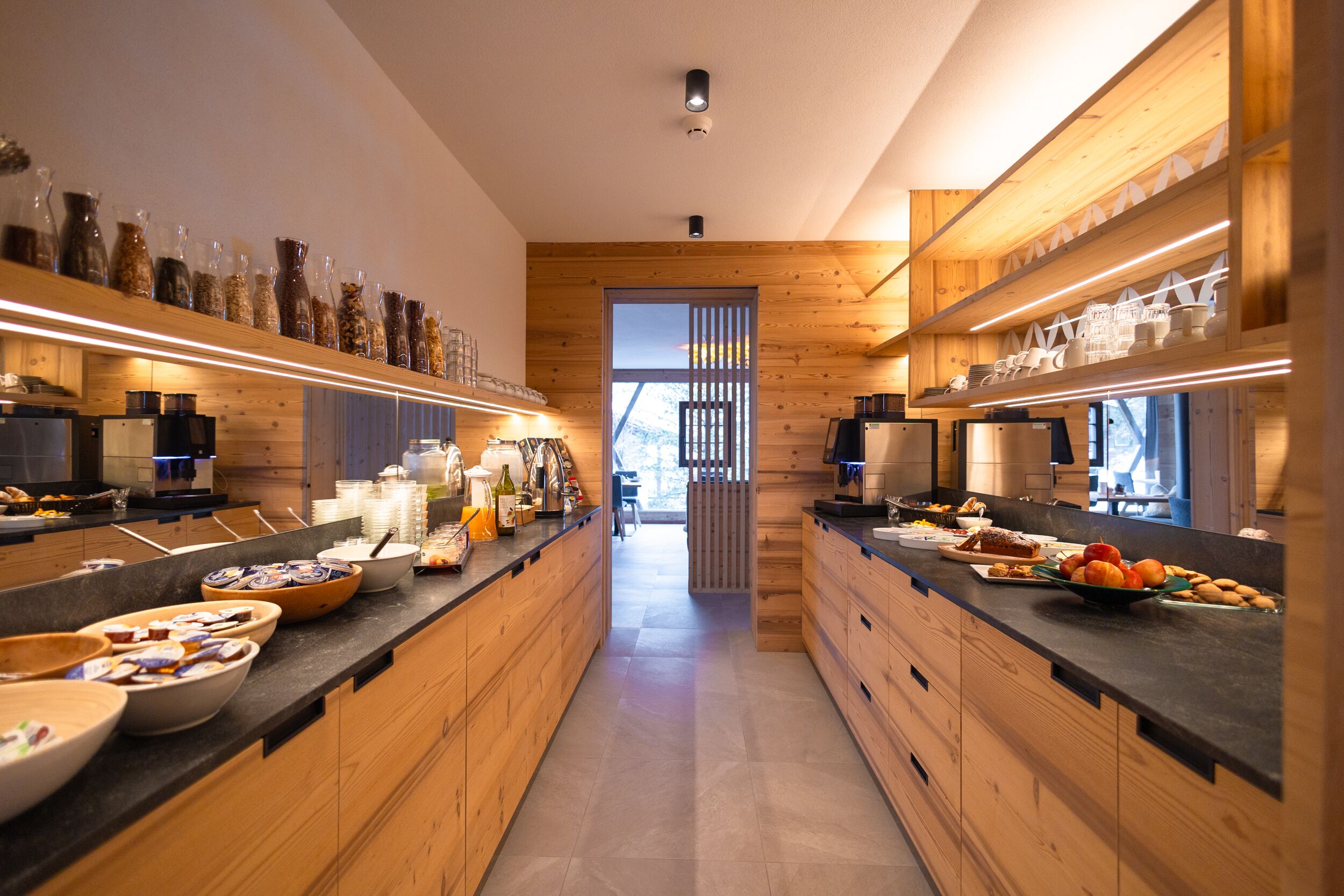Een moderne keuken met houten kastjes, zwarte aanrechtbladen en verschillende etenswaren die op planken en aanrecht staan uitgestald.
