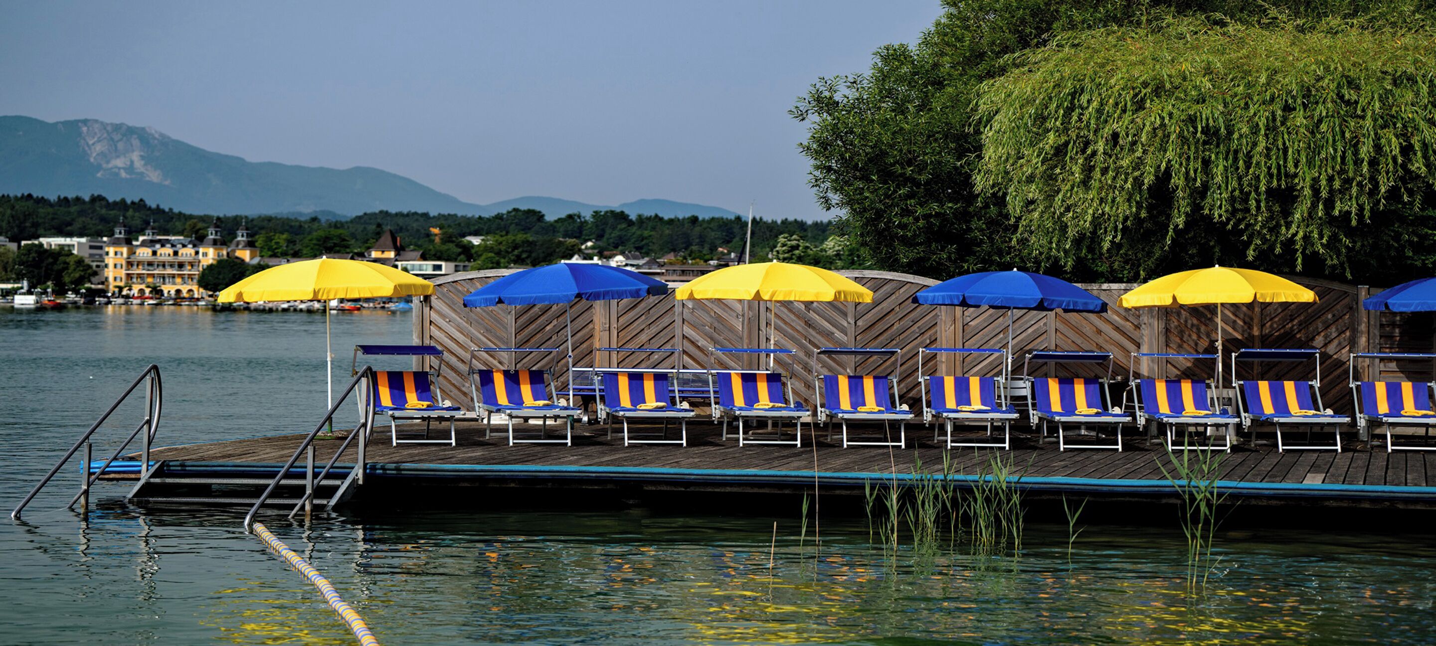 A lakeside deck with rows of blue and yellow beach chairs and umbrellas, a wooden deck, and a view of mountains in the background.