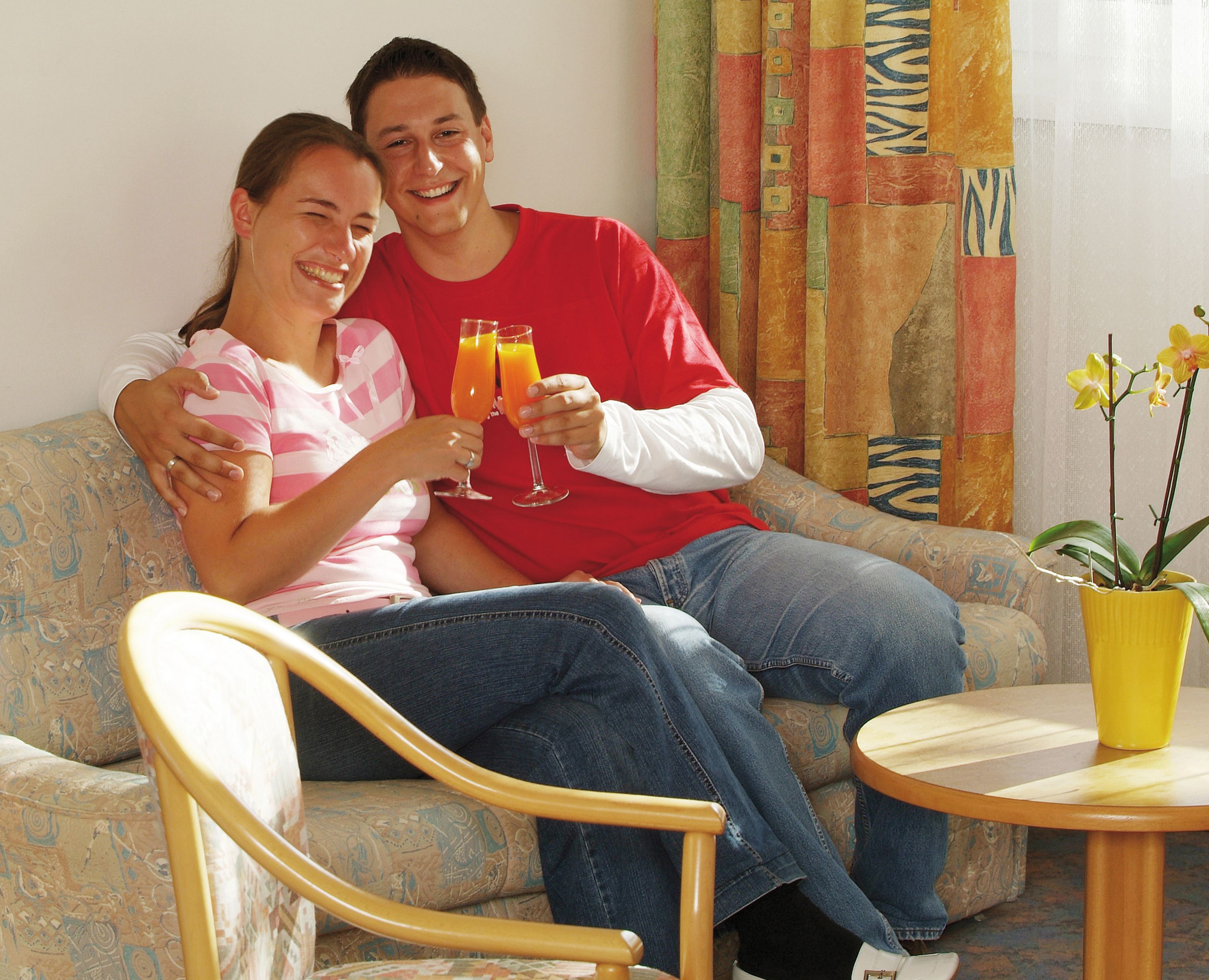 A couple sitting on a patterned sofa with a small round table and a potted plant in the foreground.