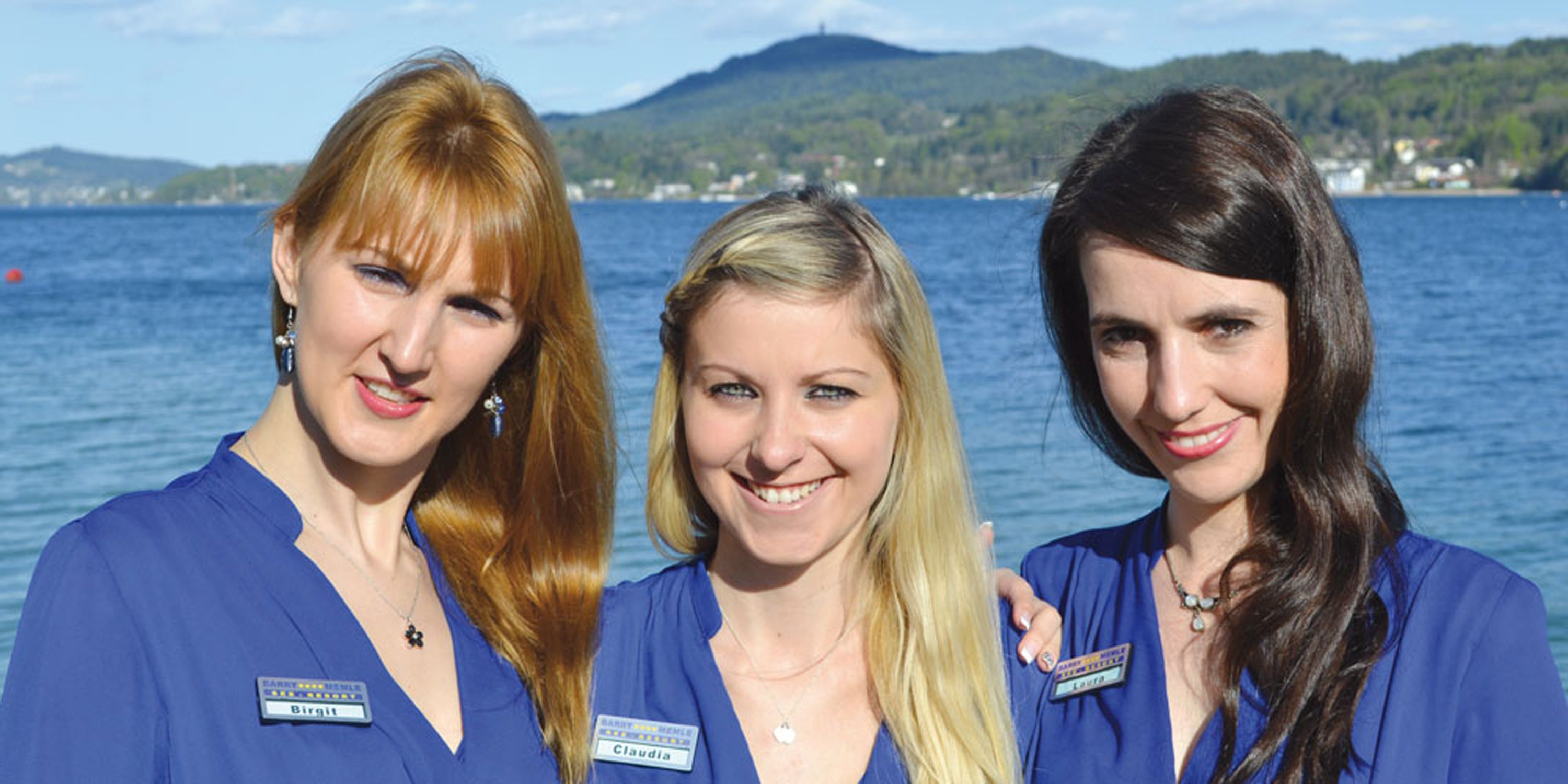 Three individuals in blue uniforms with name tags, standing by a body of water with a mountainous backdrop.