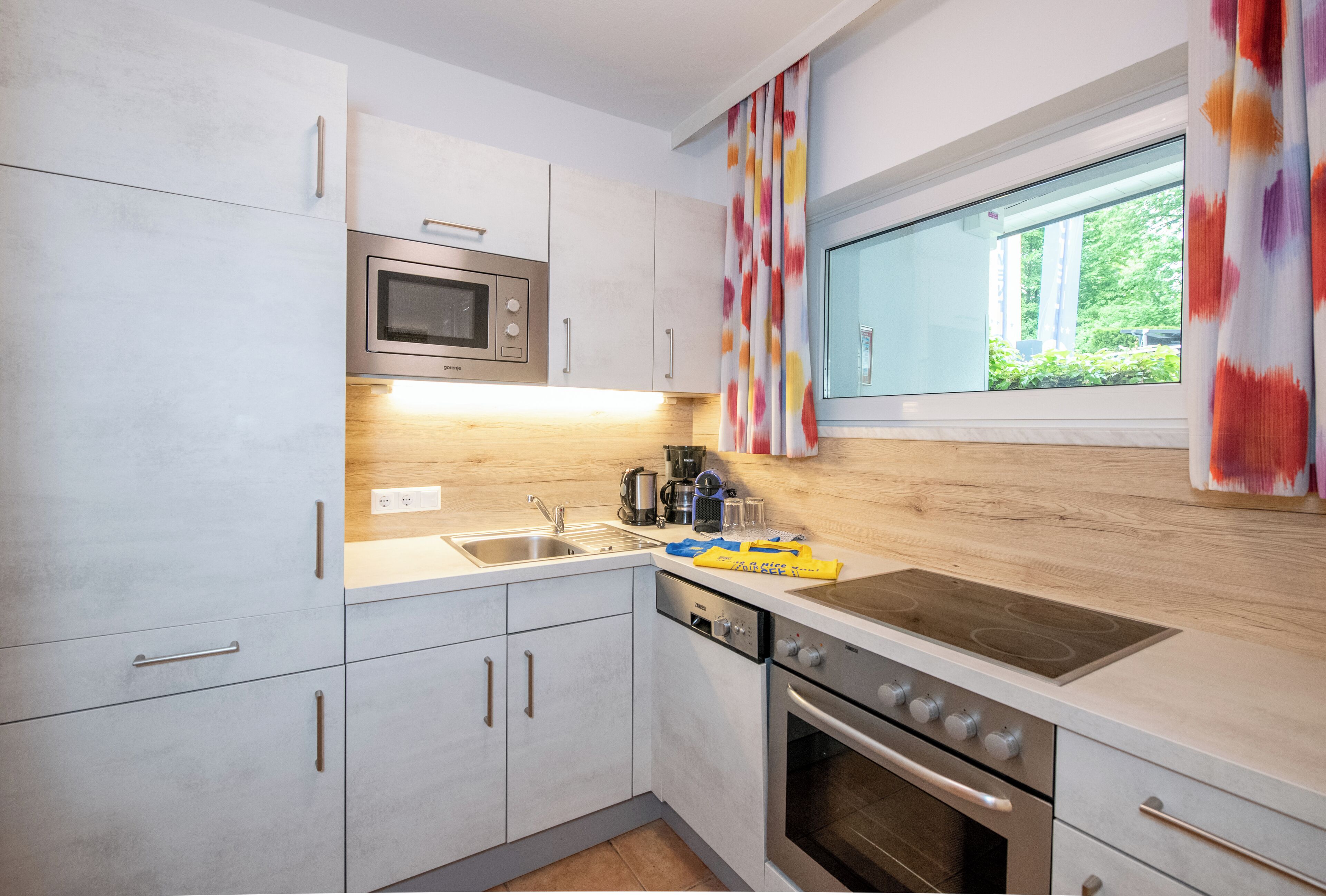 Una cocina moderna con armarios blancos, un microondas de acero inoxidable, un horno empotrado y una ventana con vistas a zonas verdes.