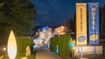 L’entrée d’un hôtel, avec un chemin menant au bâtiment, des banderoles représentant l’hôtel et les appartements, et une grande enseigne lumineuse.