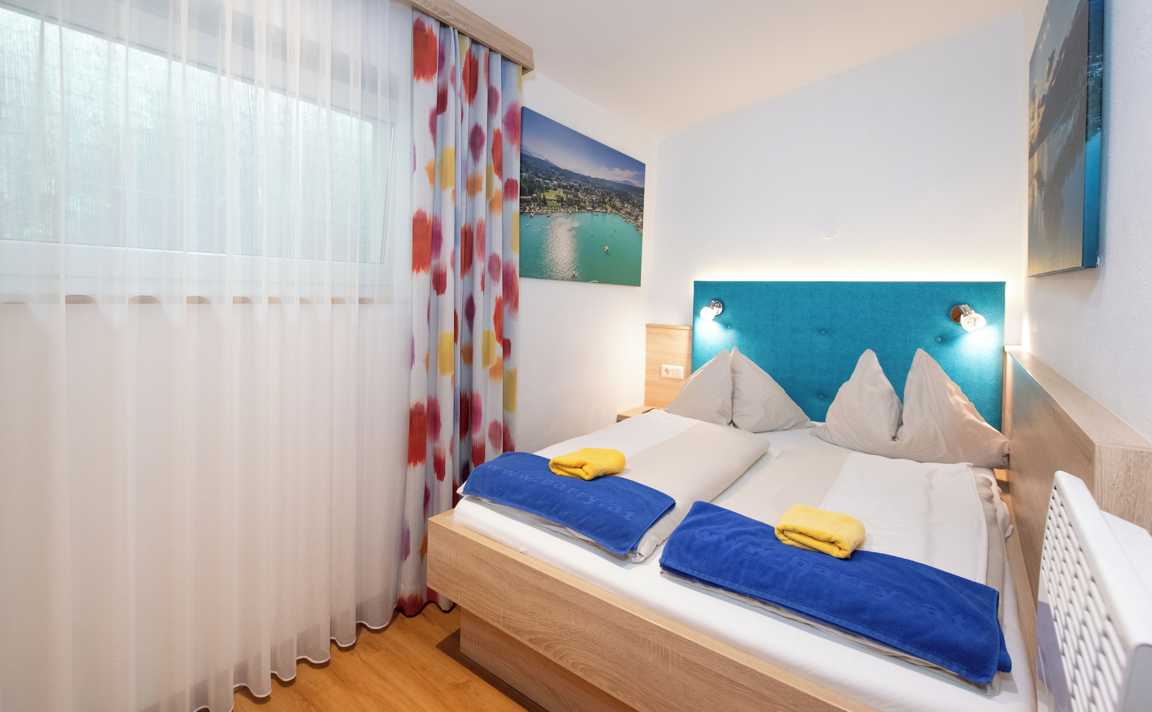 Une chambre à coucher avec un lit, une table de chevet, une fenêtre avec des rideaux légers et un tableau représentant un paysage.