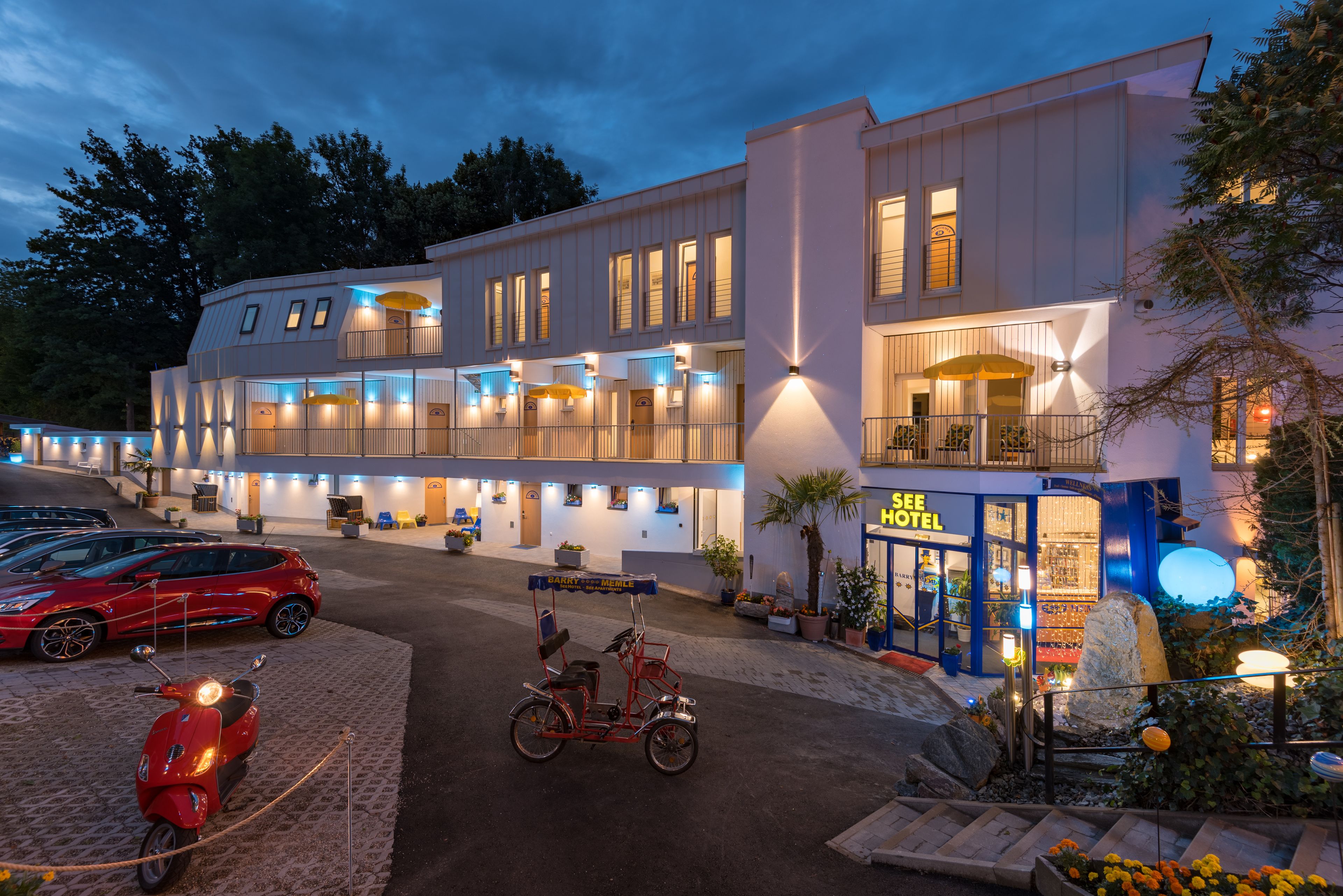 Een hotel met meerdere balkons, een zichtbare ingang en een parkeerplaats met auto's en een scooter.