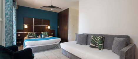 Une chambre d’hôtel moderne avec un canapé gris, un lit doté d’une tête de lit bleue, une armoire en bois foncé et un rideau bleu.