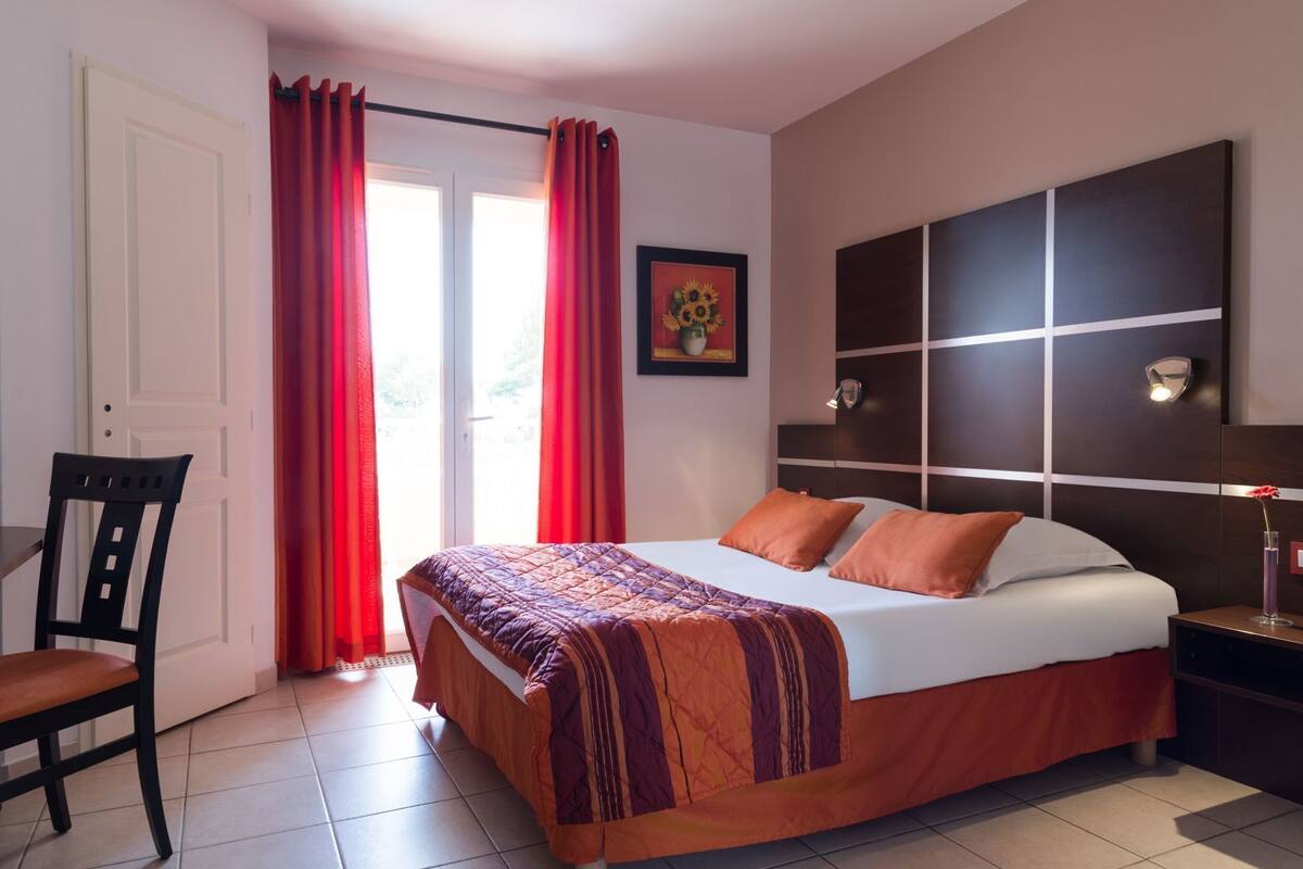 Een slaapkamer met een bed, een stoel, een nachtkastje, een wandlamp en een raam met gordijnen.