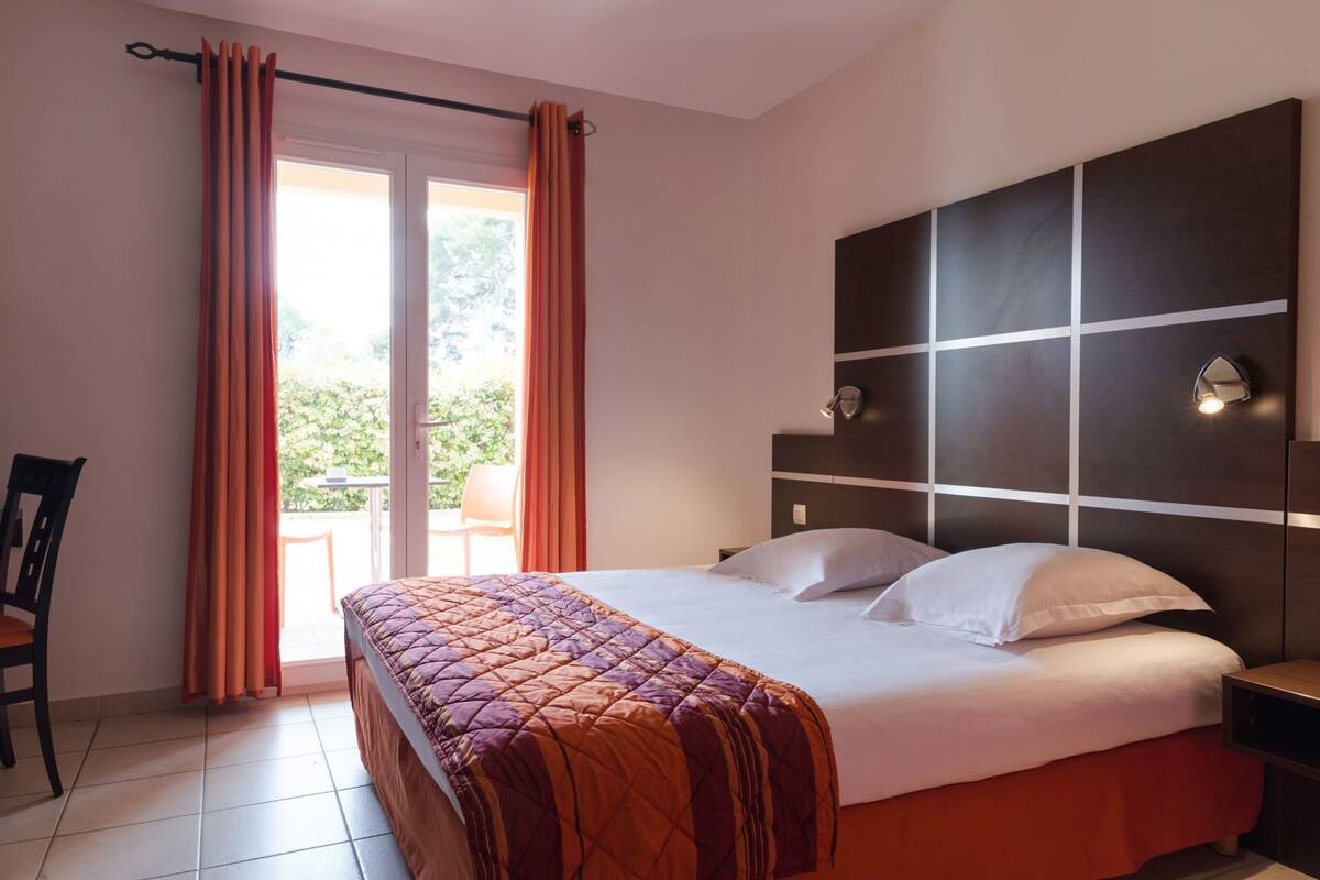 Een hotelkamer met een bed, gordijnen, een stoel, een nachtkastje en uitzicht op de omgeving.