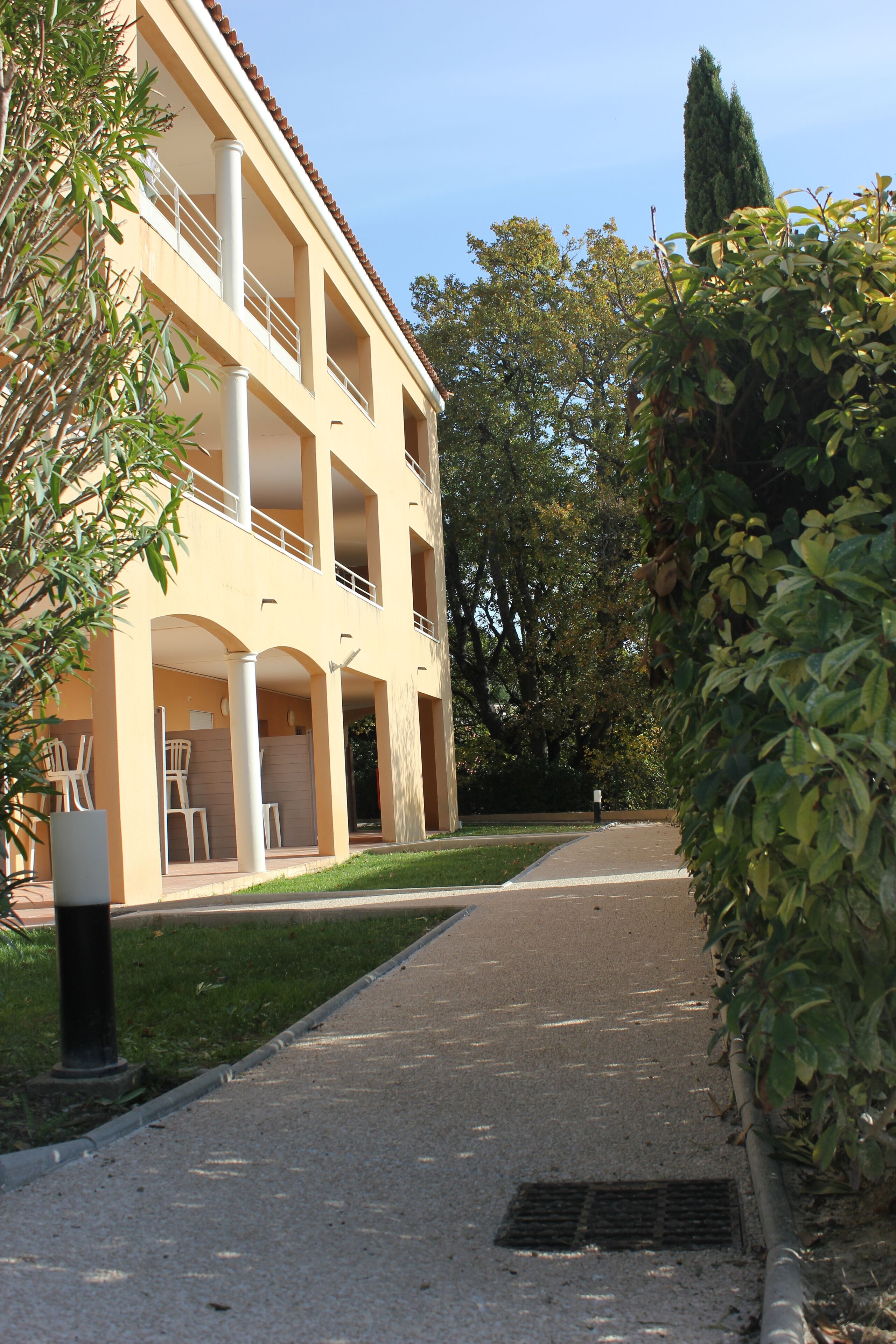 Un bâtiment à deux étages doté de balcons, d’une allée pavée et entouré de verdure.