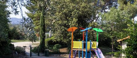 A playground with a slide and climbing structures, surrounded by trees and a gravel path.