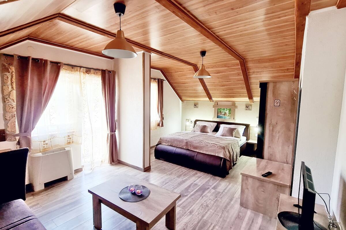 A cozy, well-lit room with a wooden ceiling, a bed with a headboard, a desk with a chair, and a television.