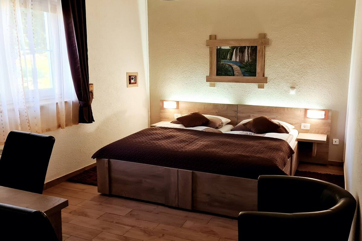 A bedroom with a bed, bedside tables, a headboard with a painting, a chair, and a window with curtains.