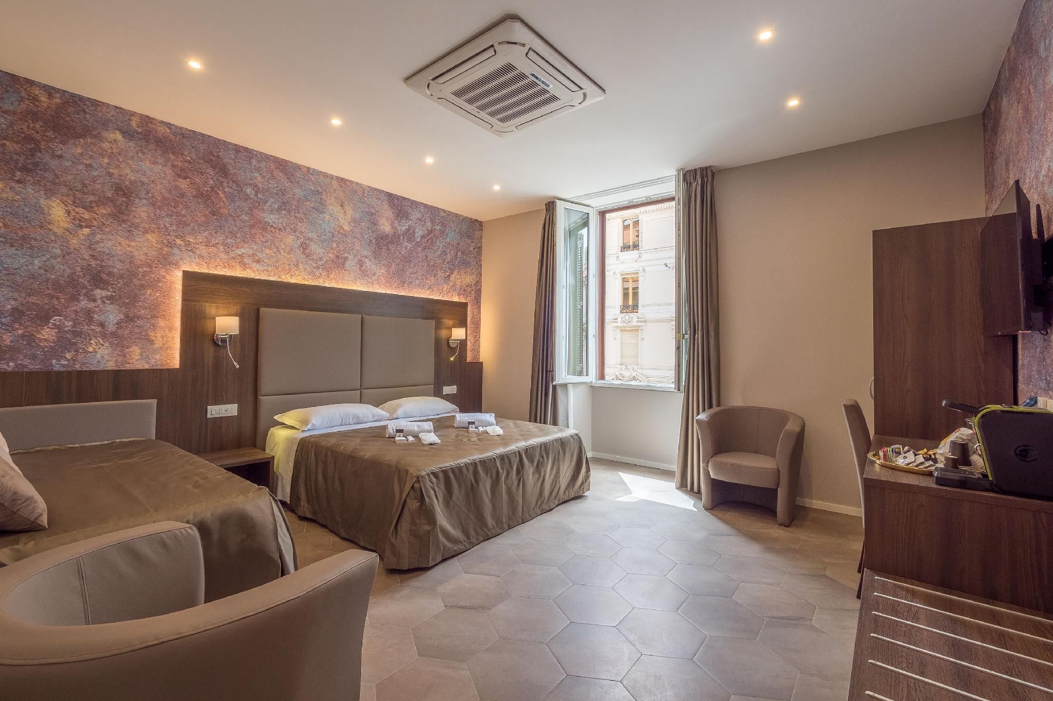 A hotel room with a large bed, two armchairs, a desk with a laptop, a window with curtains, and a textured wall.