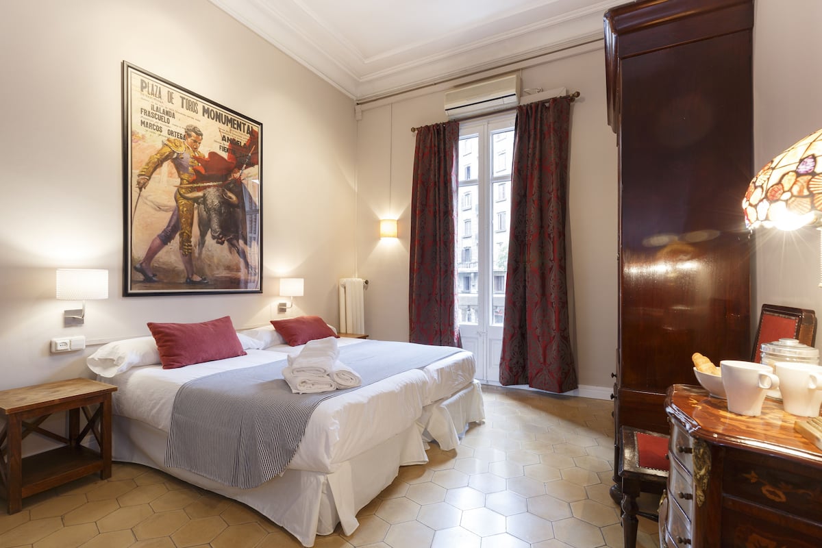 Ein Hotelzimmer mit einem großen Bett, zwei Nachttischen, einem Schminktisch, einem Kleiderschrank, einem Fenster mit Vorhängen und einer Wand-Klimaanlage.