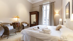 Une chambre d’hôtel avec un grand lit, une chaise en osier, une commode en bois et une fenêtre avec des rideaux.