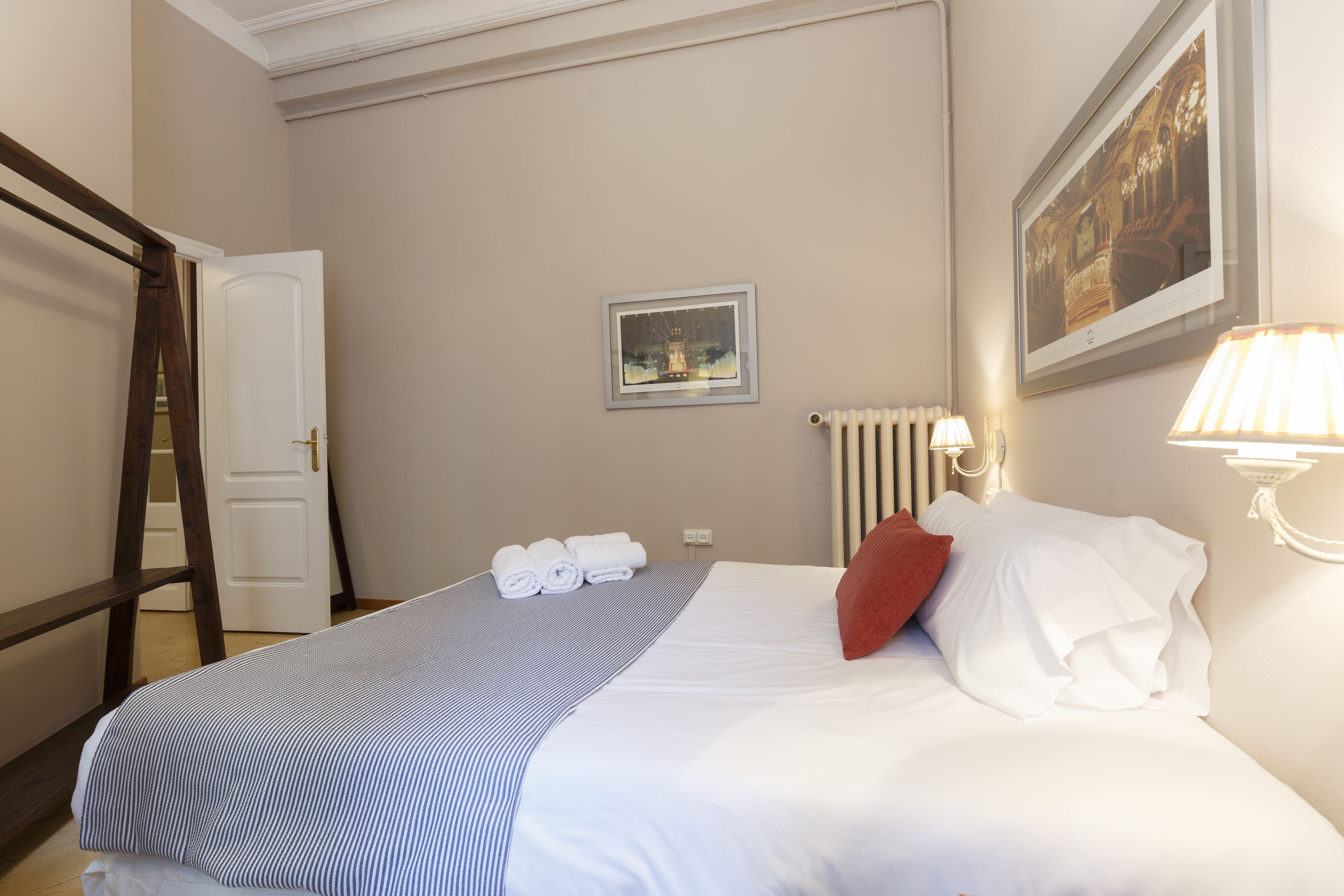Een netjes opgemaakt bed met witte en gestreepte beddengoed, een rood kussen en een ingelijst fotokader aan de muur.